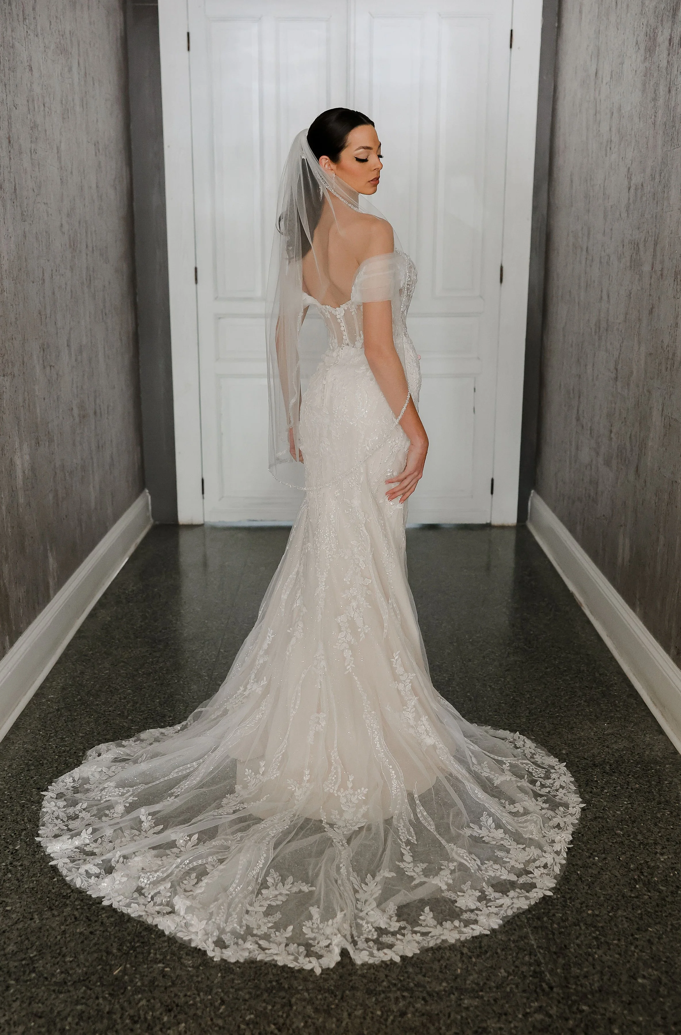 A bride in an elegant, strapless, off-the-shoulder lace wedding gown with a long train and a veil, standing in a hallway with gray walls and closed white doors behind her.