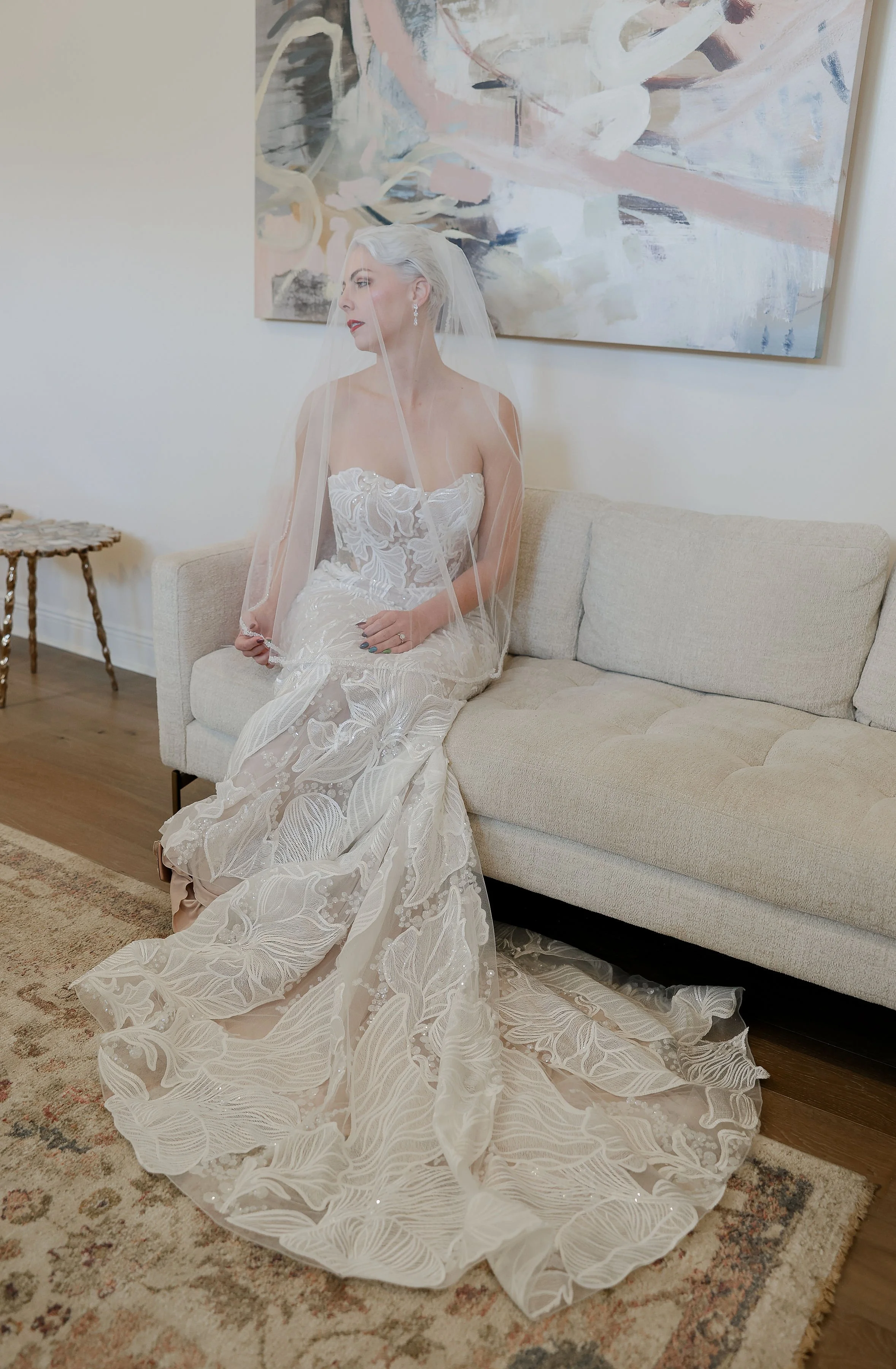 A woman dressed in a white wedding gown sitting on a beige sofa with her veil covering part of her face. She has silver hair styled in waves, red lipstick, and earrings. A large abstract painting is on the wall behind her, and a small wooden side tab
