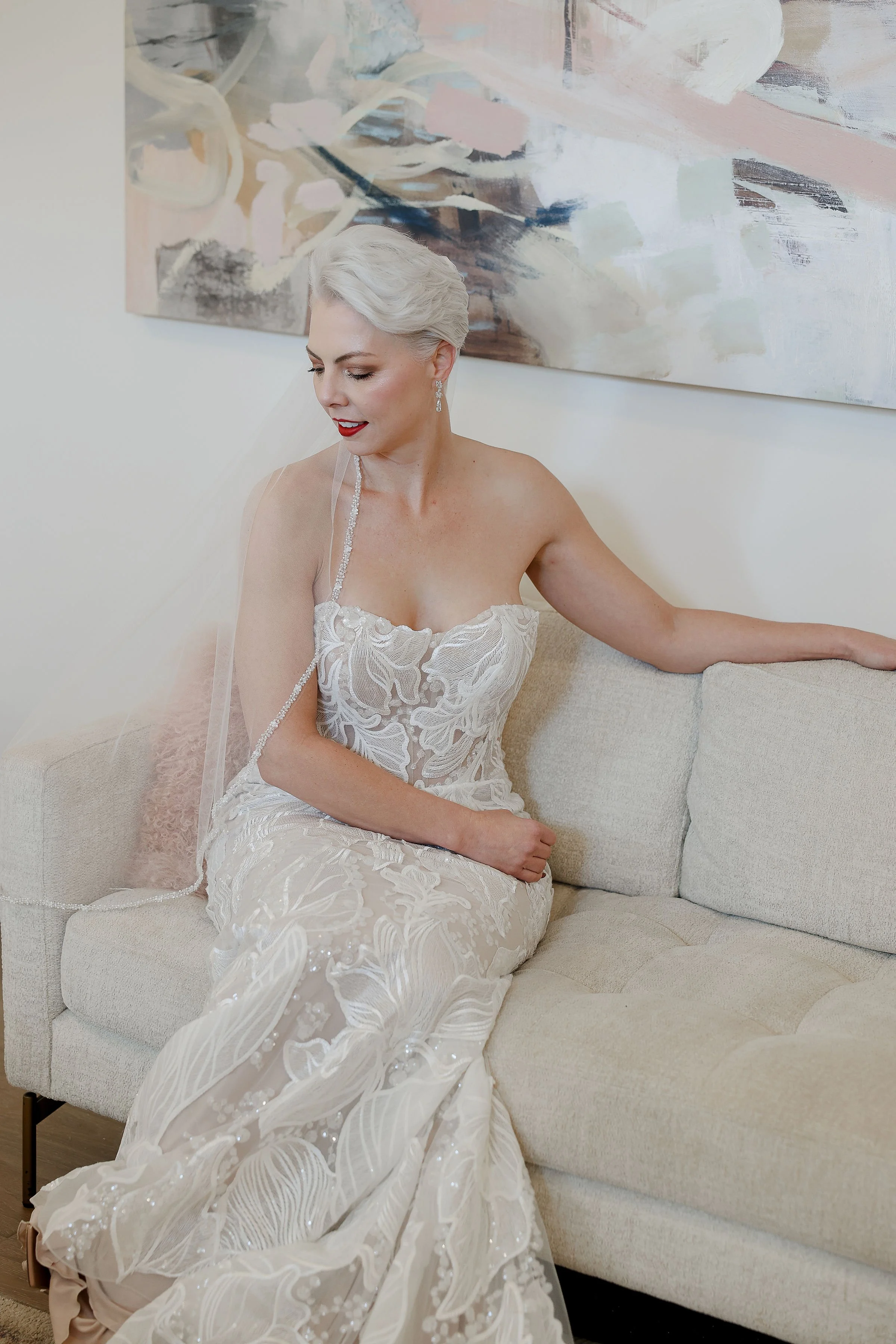 A bride with platinum blonde hair sitting on a beige sofa, wearing a white lace wedding dress with embroidery, earrings, and a veil. A large abstract painting hangs on the wall behind her.