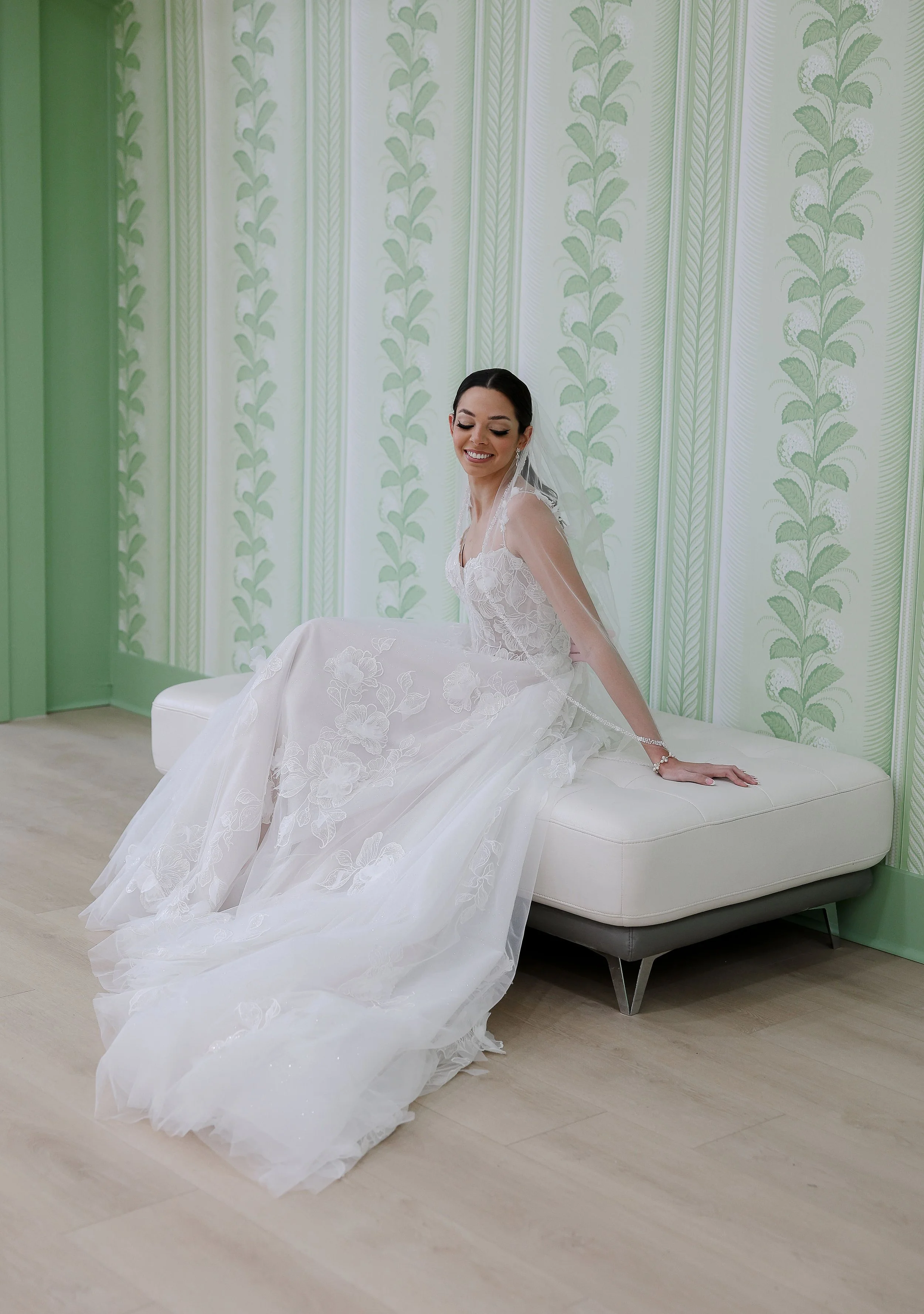 A smiling bride in a white wedding gown sitting on a white bench in a room with green and white patterned curtains.