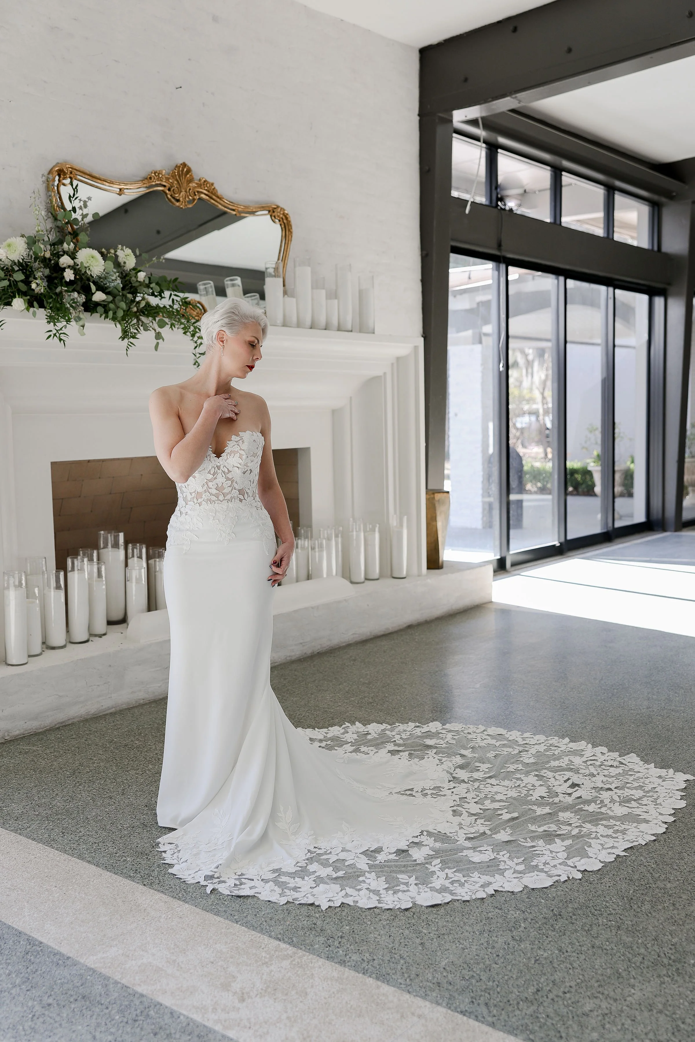 A woman in a white wedding dress standing in a modern interior with large windows, a fireplace decorated with candles and flowers, and a carpet with flower petal designs.