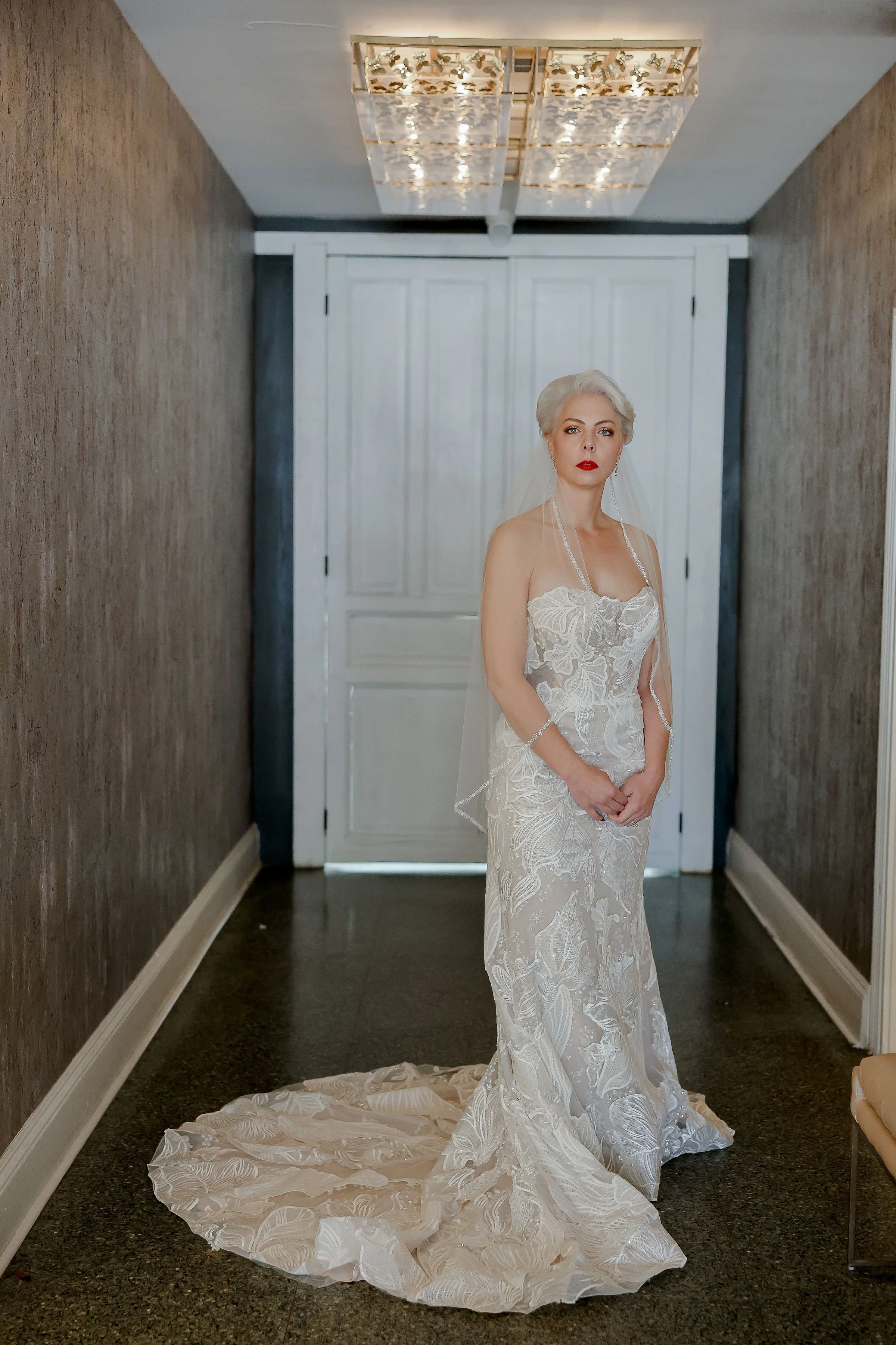 A woman in a wedding dress standing in a hallway with white double doors and chandeliers above.
