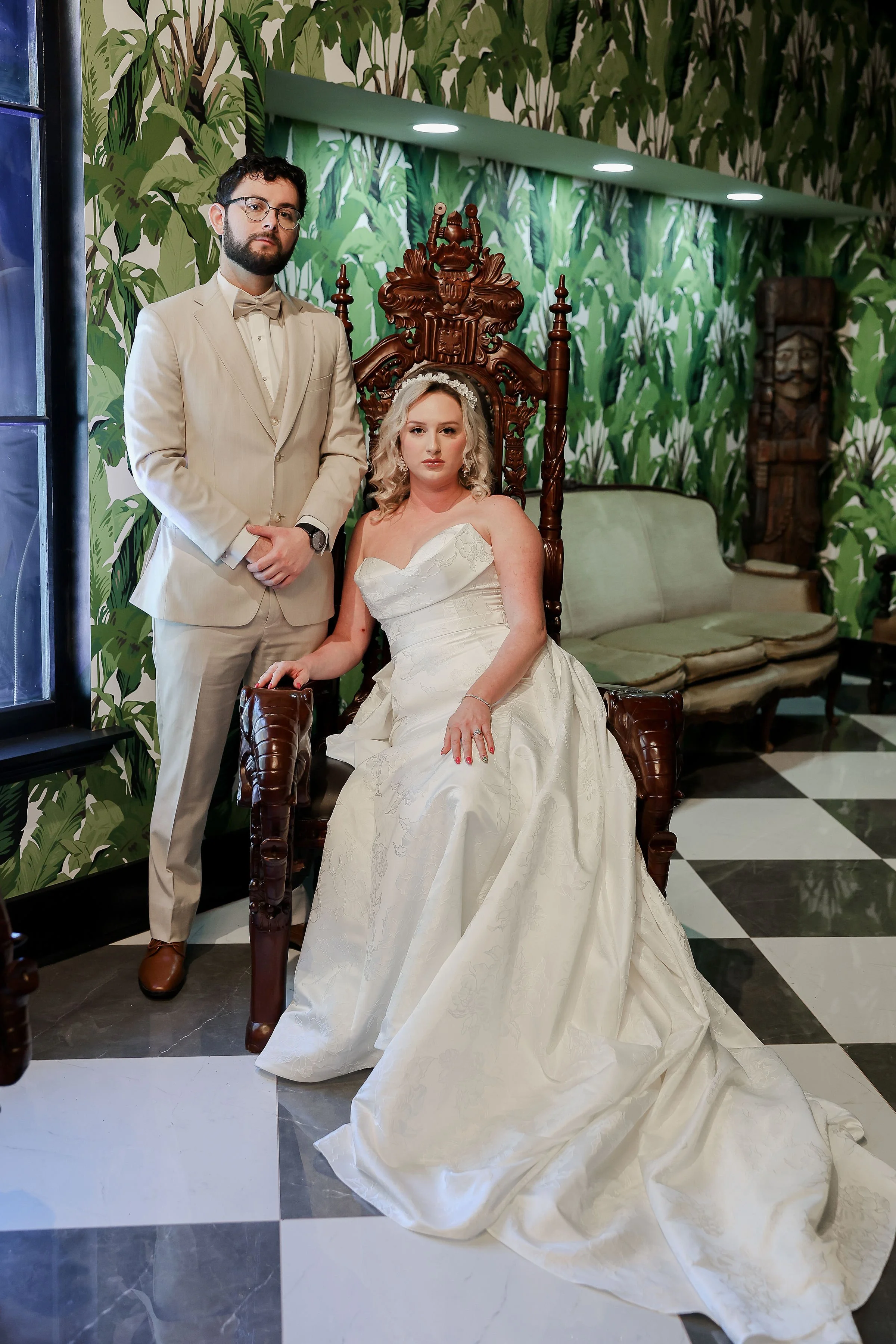 A man with glasses, a beard, and a light beige suit standing next to a seated woman in a white wedding gown with a full skirt and strapless neckline. They are in a room with leafy green wallpaper and wooden African art decor.