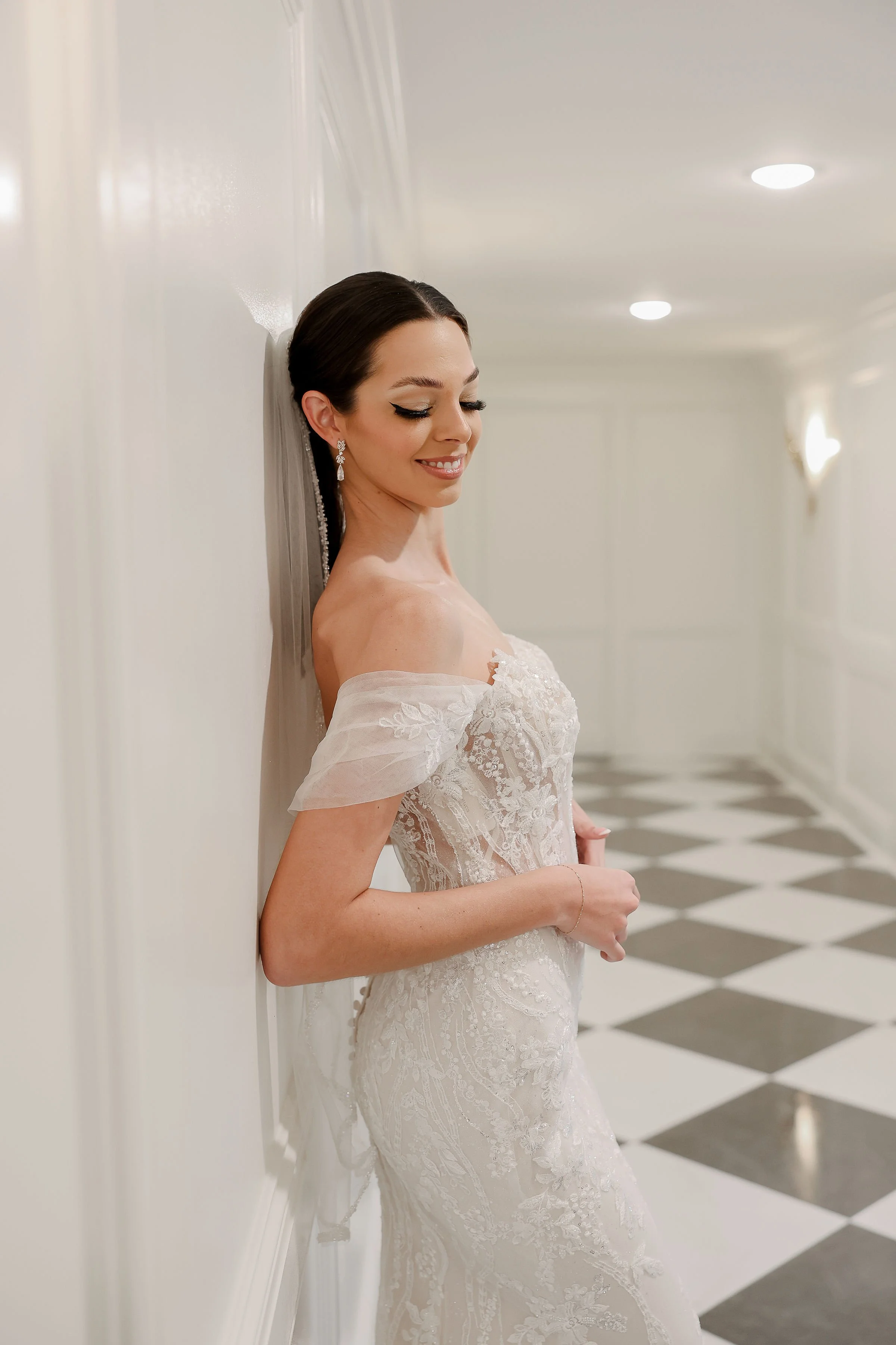 A bride with dark hair in an elegant updo, wearing diamond earrings and a strapless lace wedding gown, stands against a white wall in a corridor with checkered black-and-white flooring, smiling softly with eyes closed.