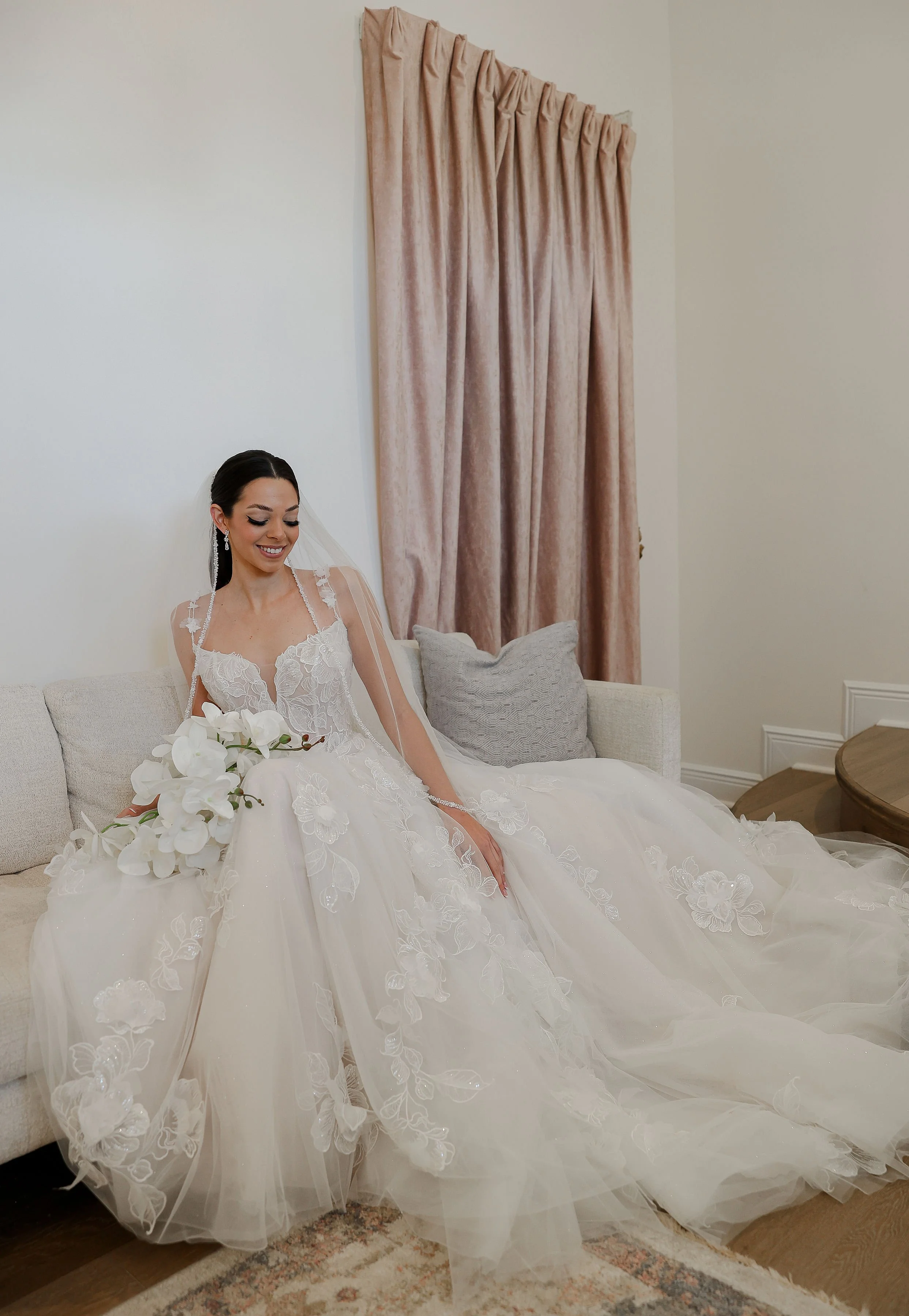 A bride sitting on a light-colored sofa in a wedding dress, holding a bouquet of white orchids, smiling softly in a room with beige curtains and a patterned rug.