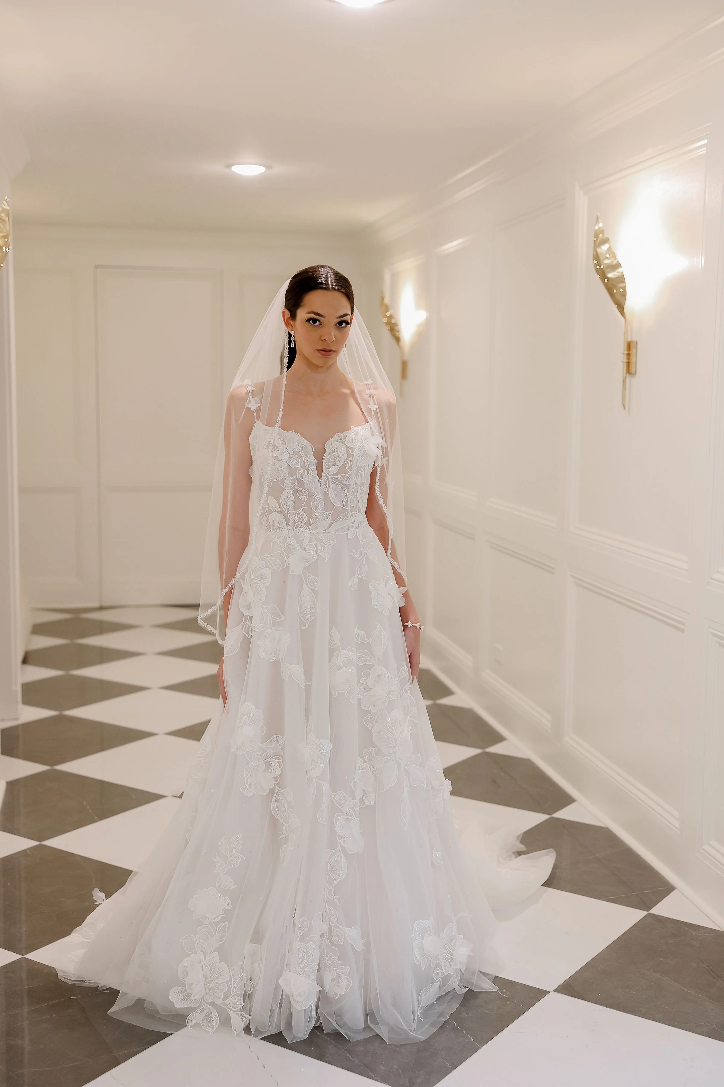 A woman in a white wedding dress and veil standing in a well-lit, elegant corridor with white paneled walls and gold wall sconces.