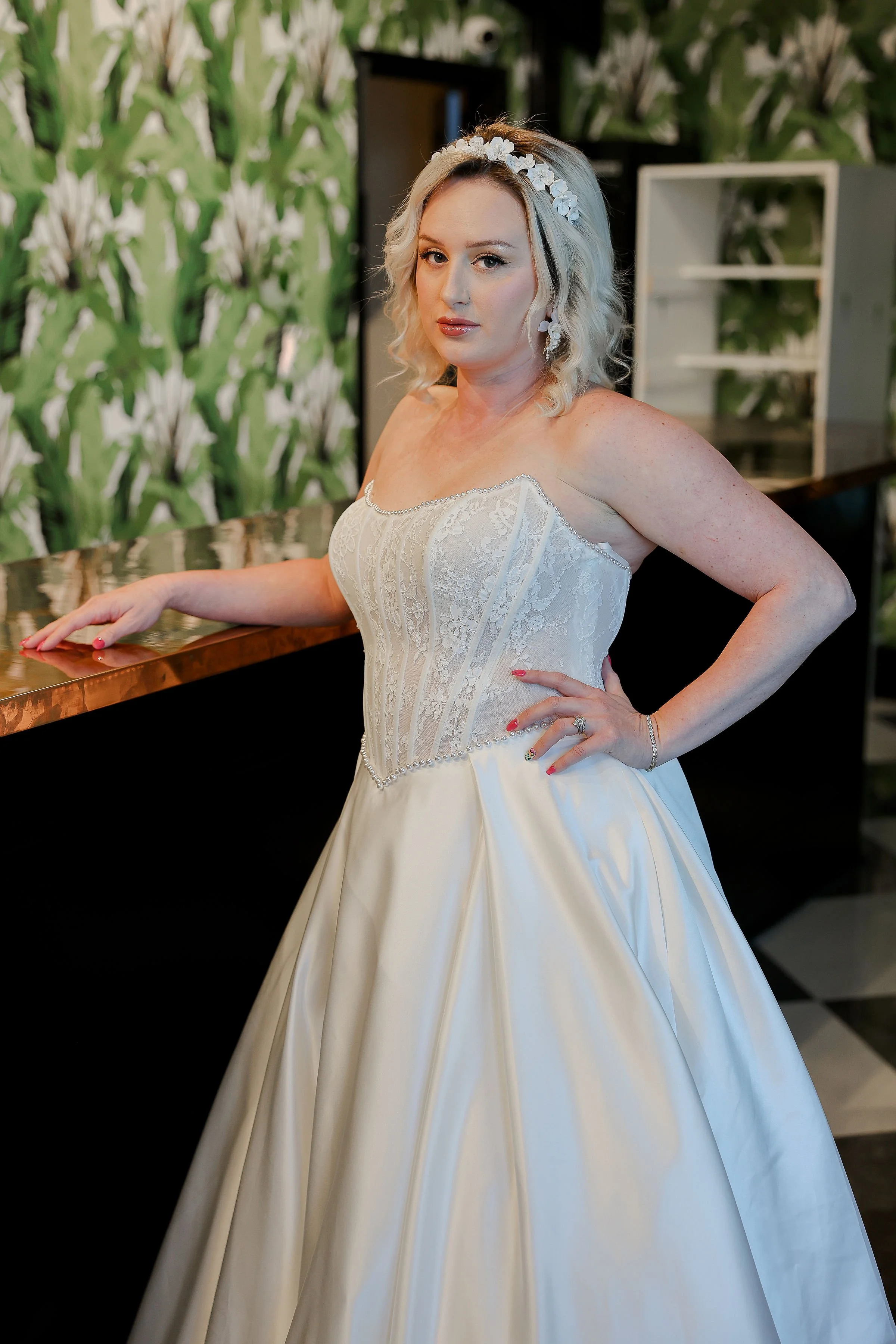 A woman in a wedding dress standing indoors with a floral headband, in front of a leafy green wallpaper background, leaning on a copper-colored bar with her hand relaxed on the surface, looking at the camera.