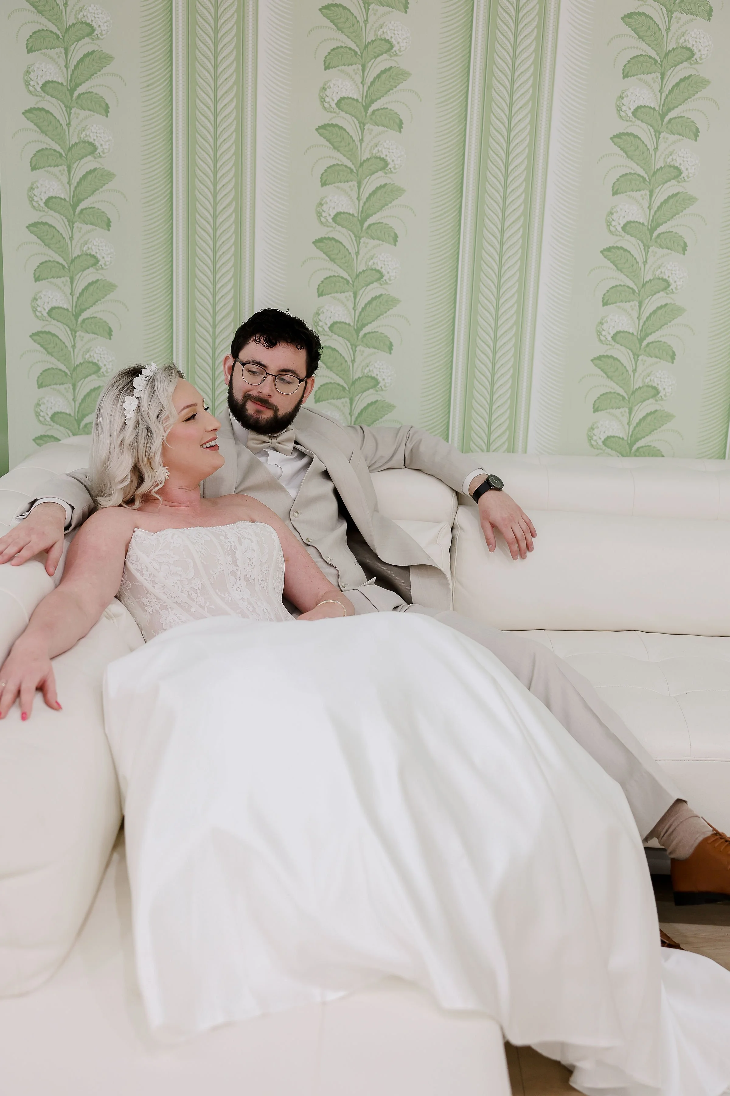 A couple sitting on a white sofa in front of a green patterned wallpaper. The woman has blonde hair, a floral headband, and a strapless white dress. The man has dark hair, glasses, a beard, and is wearing a beige suit with a bow tie. They are smiling