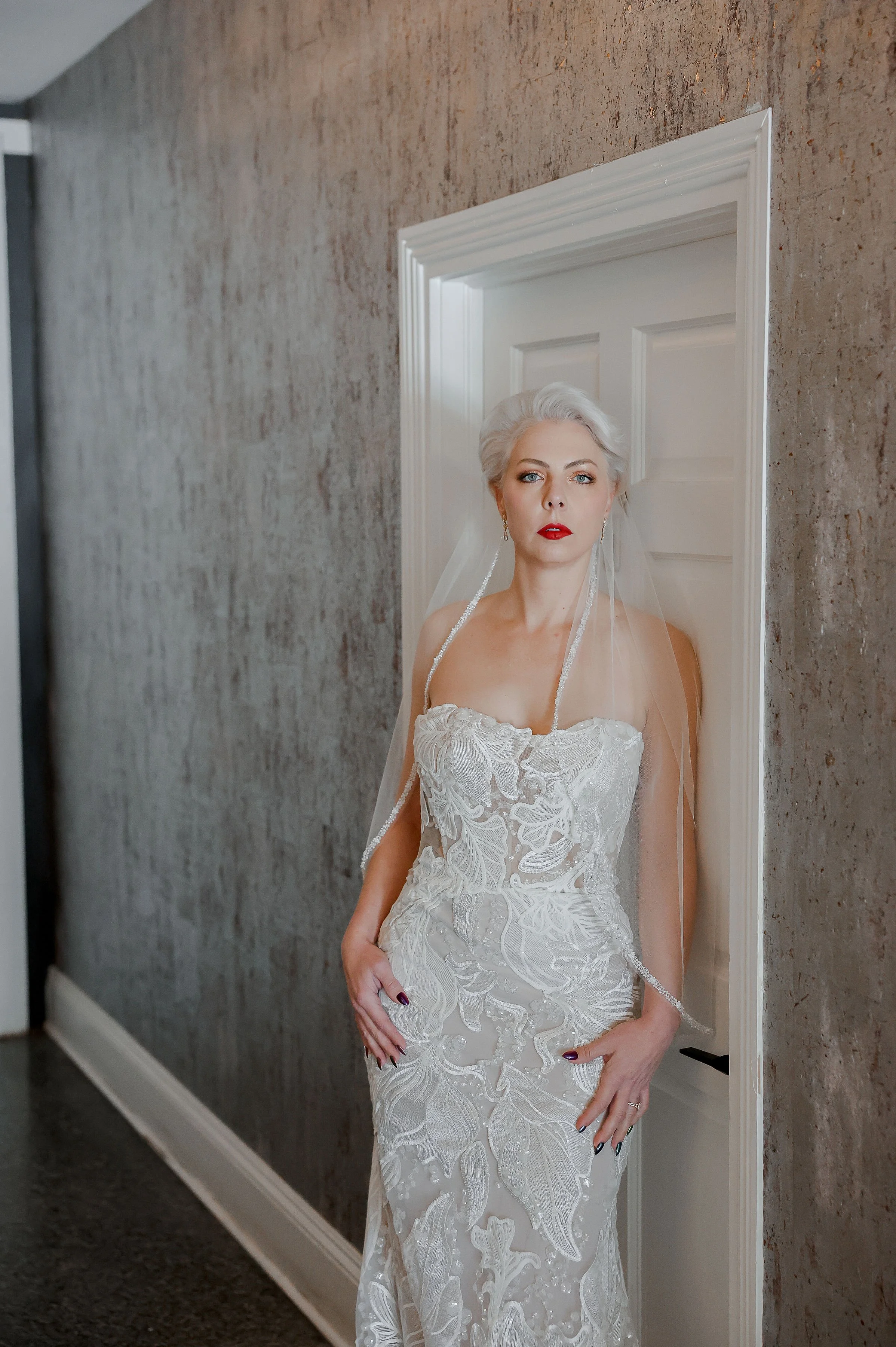 A woman in a white wedding dress and matching veil standing in front of a mirror framed in white, with a grayish textured wall behind her.