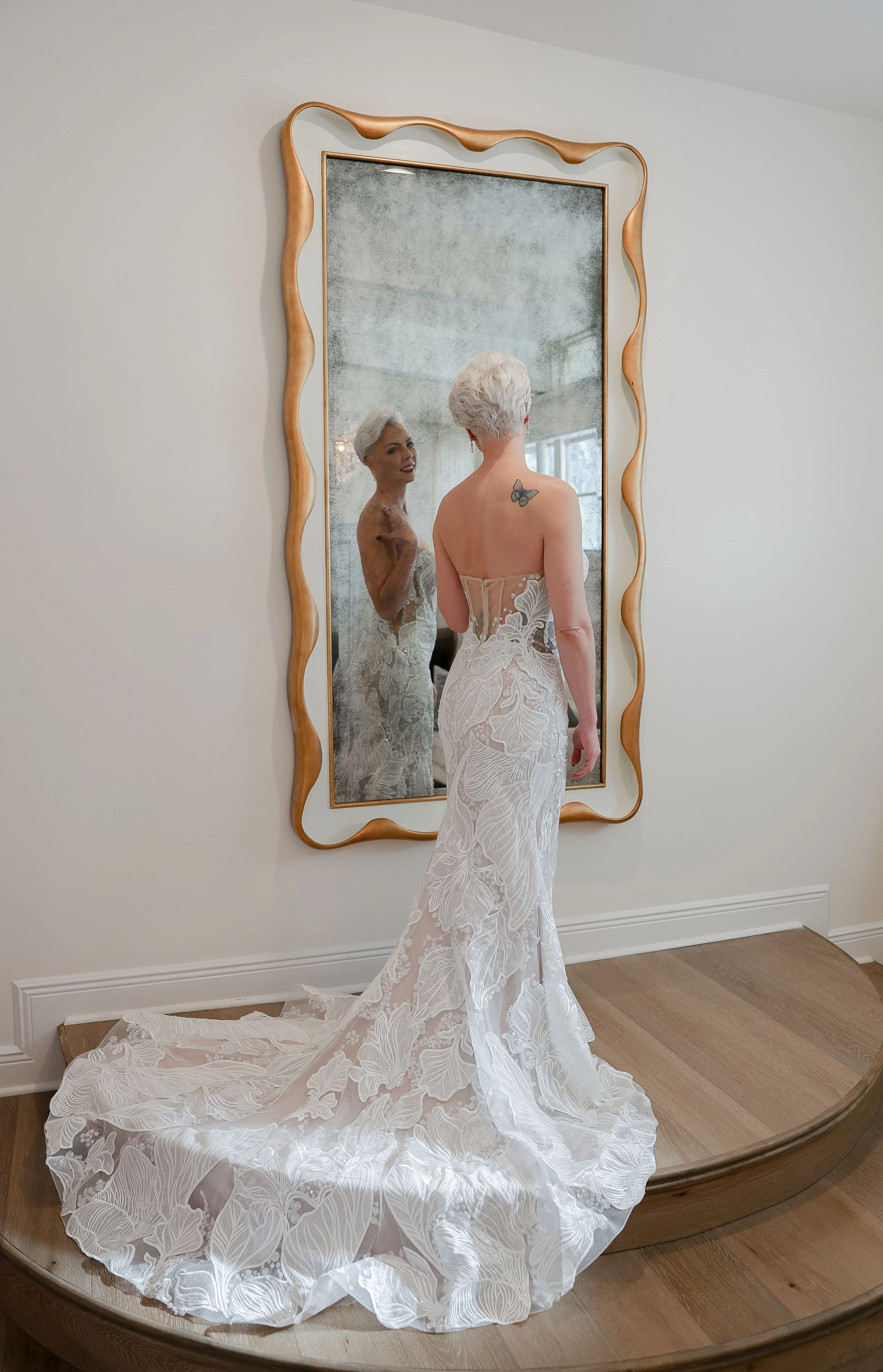 A woman with short white hair wearing a white lace wedding dress stands in front of a large mirror with a wavy wooden frame, looking at her reflection.