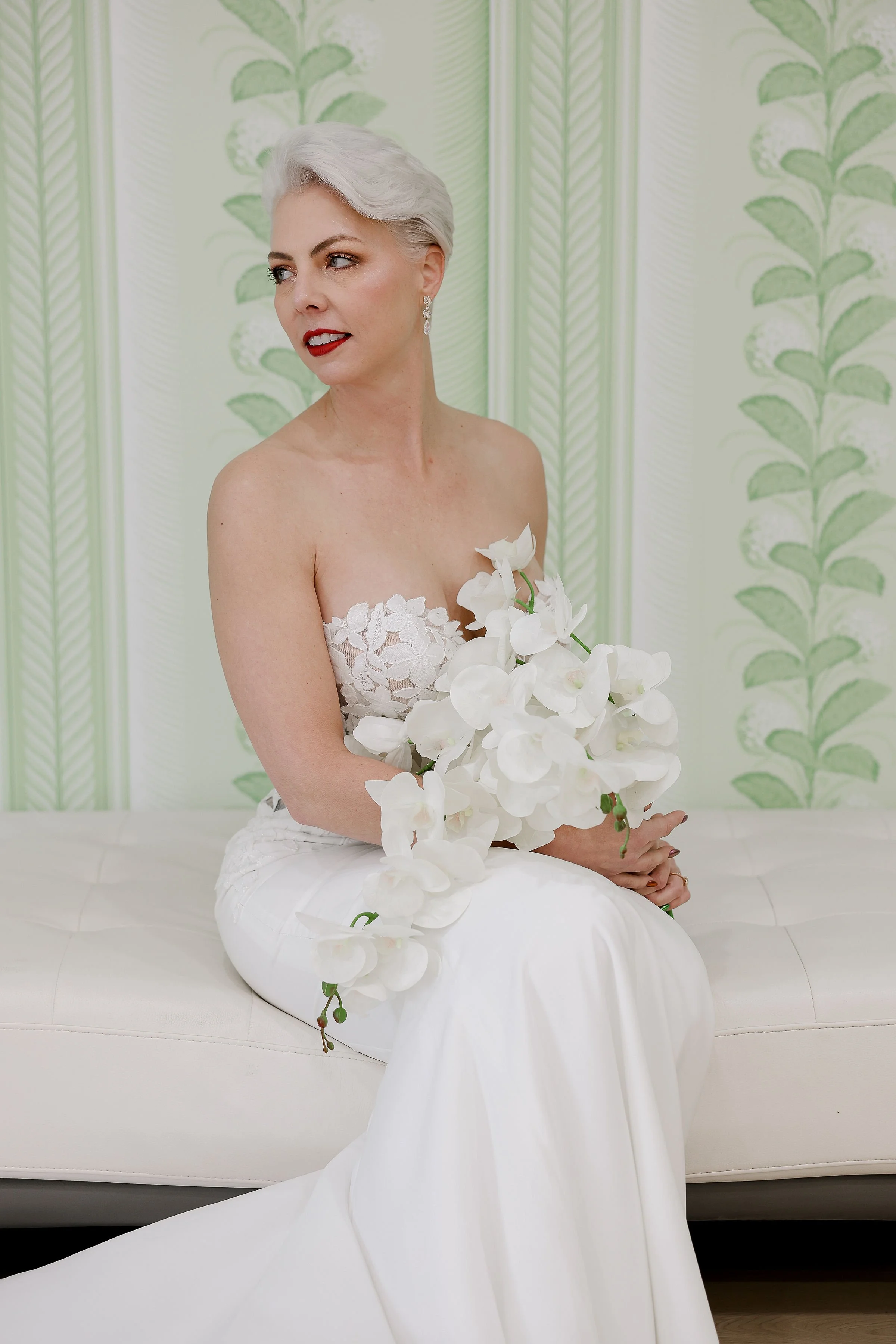 A woman with short silver hair, red lipstick, and earrings, wearing a white lace dress, sits on a white sofa, holding white orchids, with green patterned wallpaper in the background.