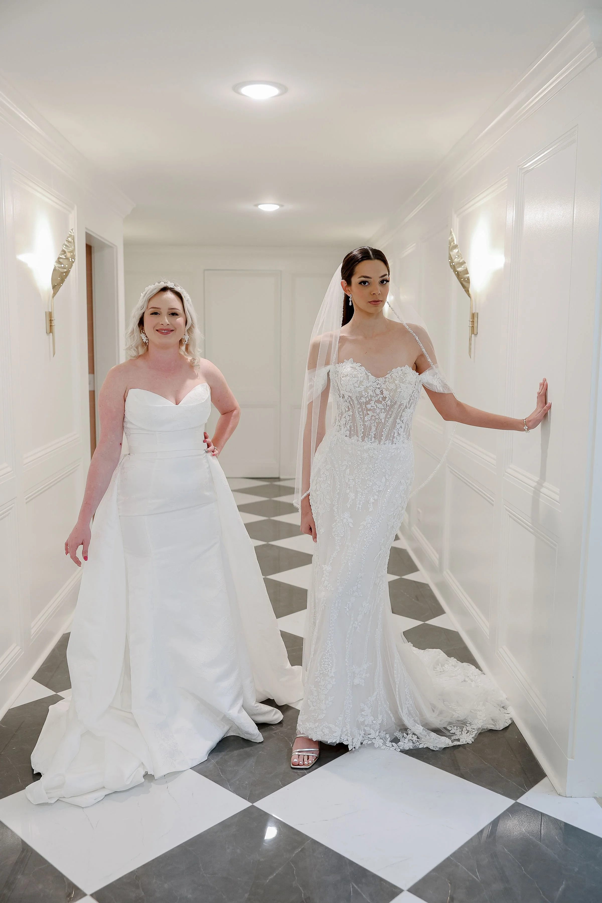 Two women in white wedding dresses standing in a hallway with white walls and black-and-white checkered floor. One woman is smiling, and the other has a neutral expression. The woman on the right is wearing a lace wedding gown with off-the-shoulder s