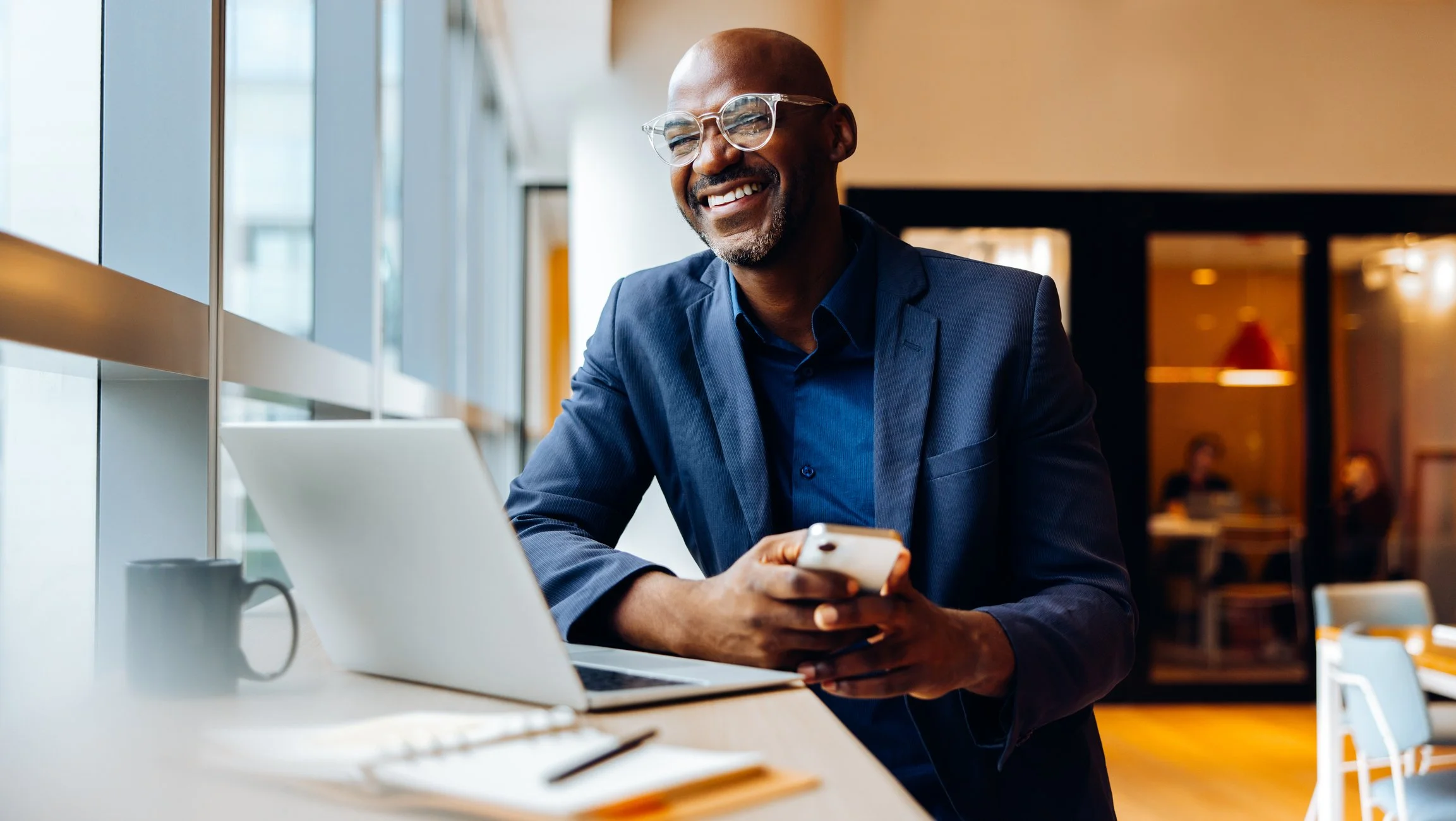 Smiling man wearing glasses in a navy blazer sitting at a table with a laptop, looking at his phone in a modern café or office space.