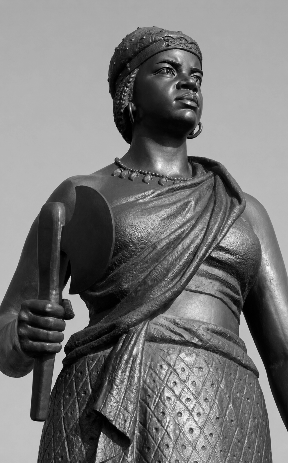 A black and white photograph of a bronze statue of a woman with traditional attire, holding a spear, with an intense and proud expression, against a plain sky background.