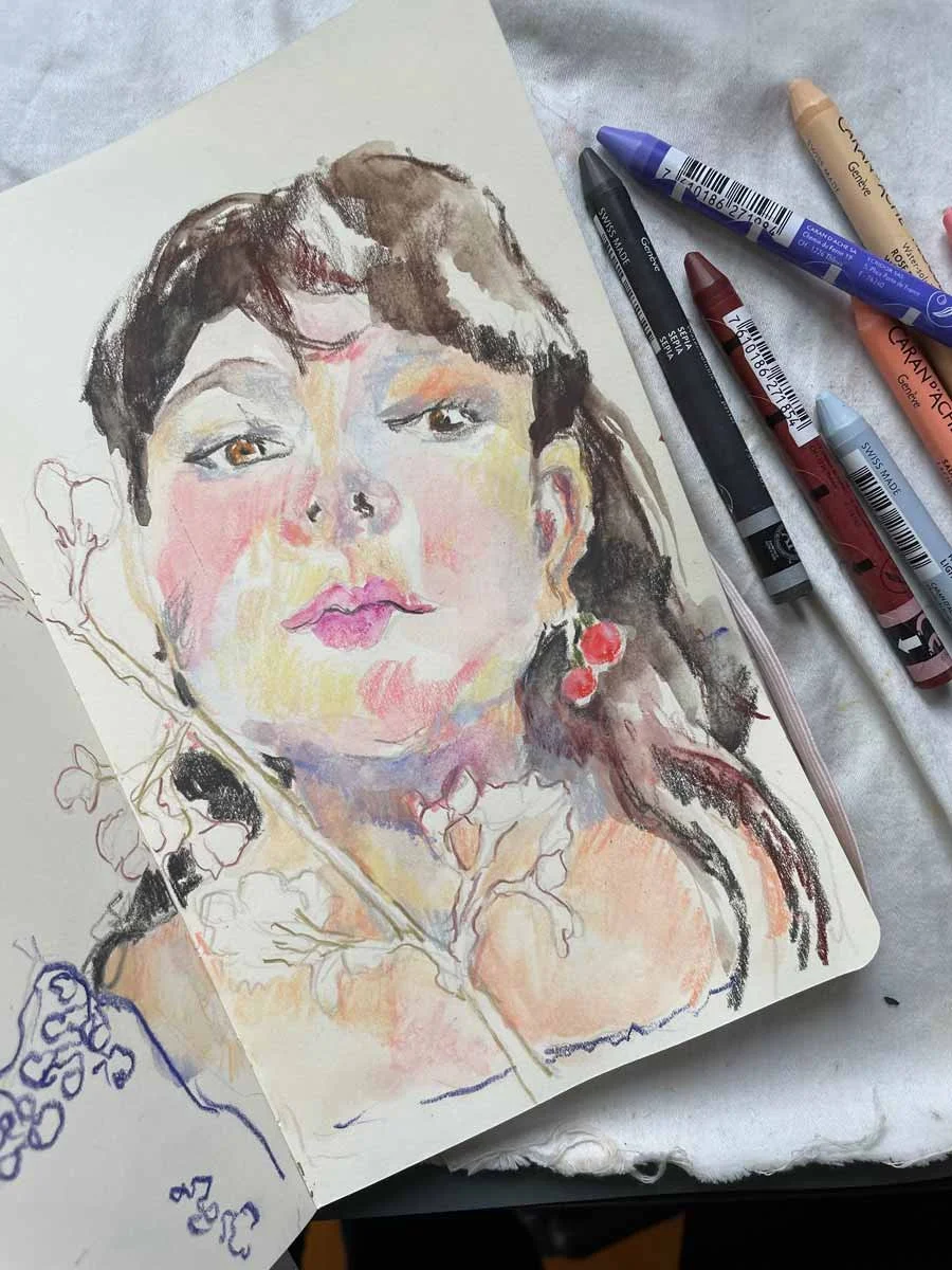 A watercolor portrait of a woman with dark hair, pink lips, and earrings, partially sketched on paper with colored markers and pencils nearby.