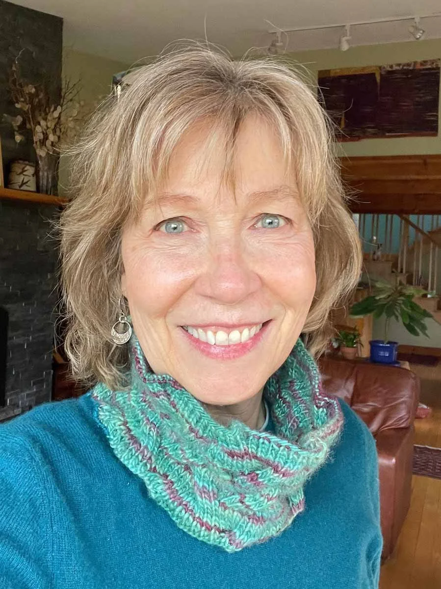 A smiling middle-aged woman with shoulder-length blonde hair, wearing a blue sweater and a multicolored knitted neck warmer, standing in a cozy living room with plants, wooden decor, and a fireplace in the background.