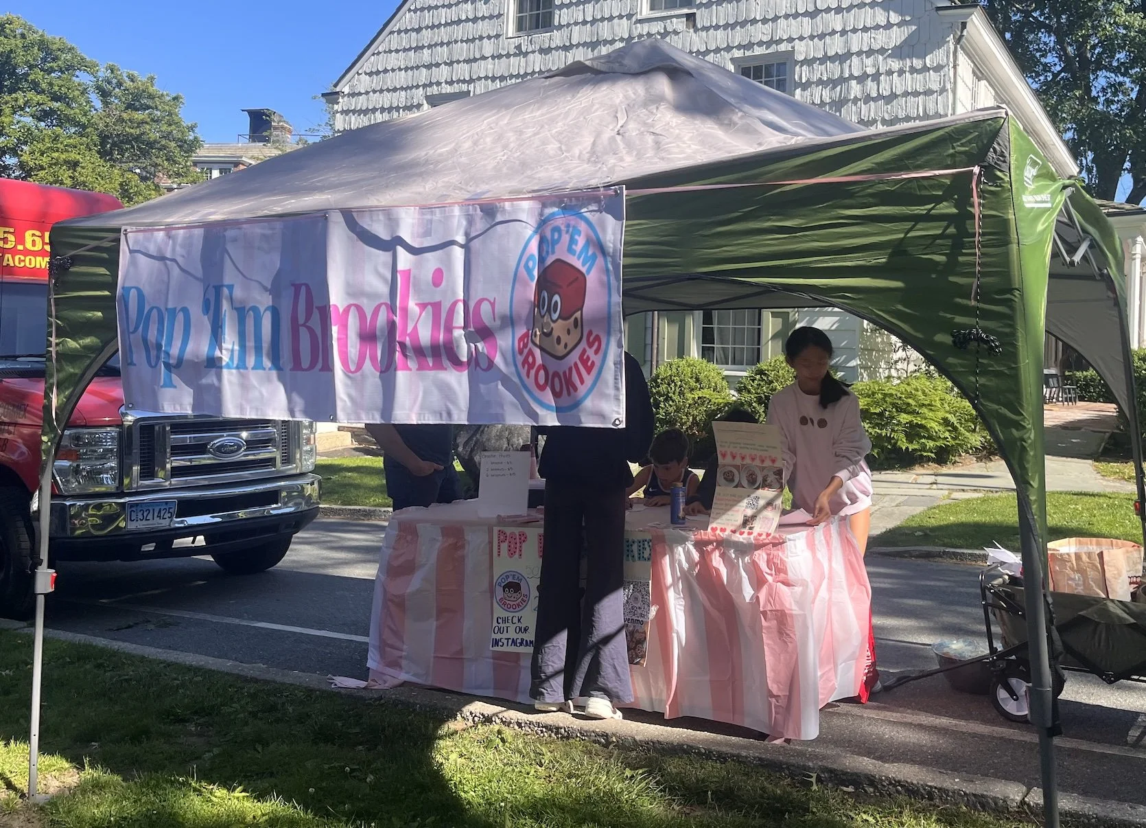 A street fair booth for 'Pop 'Em Brookies' with a pink and white striped tablecloth, a person in a light pink hoodie, a child, and a man behind the booth. There is a banner displaying the brand's name and logo, and a menu on the table.