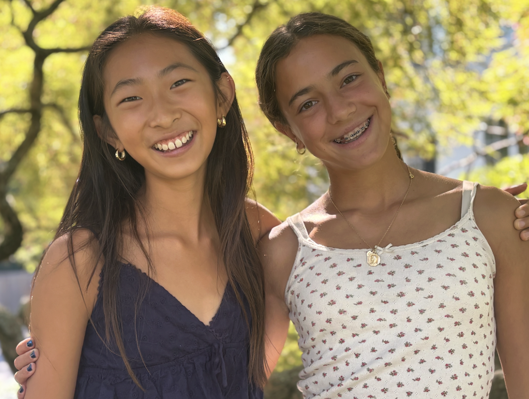 Two young women smiling, standing close together outdoors with trees in the background.