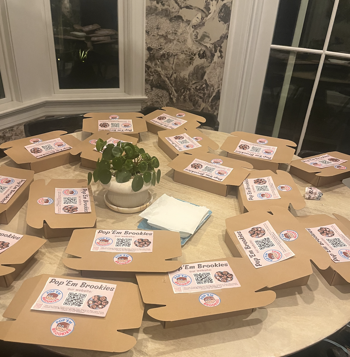 Several cardboard packaging boxes laid out on a dining table, each labeled with a sticker for 'Pop 'Em Brookies' showing a website and QR code.