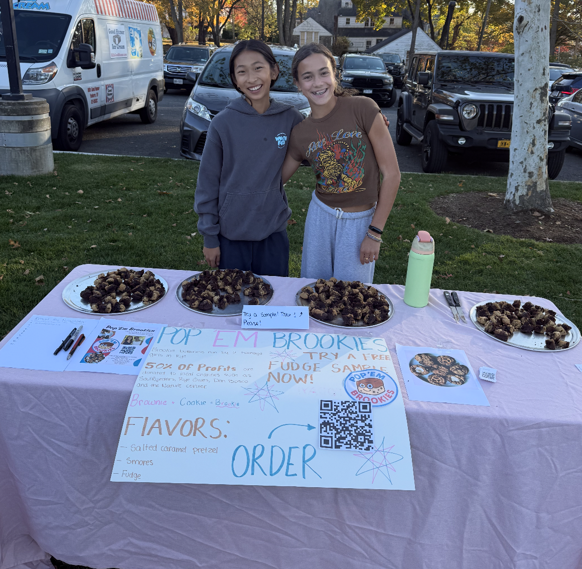 Two young girls smiling at a bake sale table with plates of brookie treats, a water bottle, and signs advertising flavors and ordering instructions for Pop 'Em Brookies.