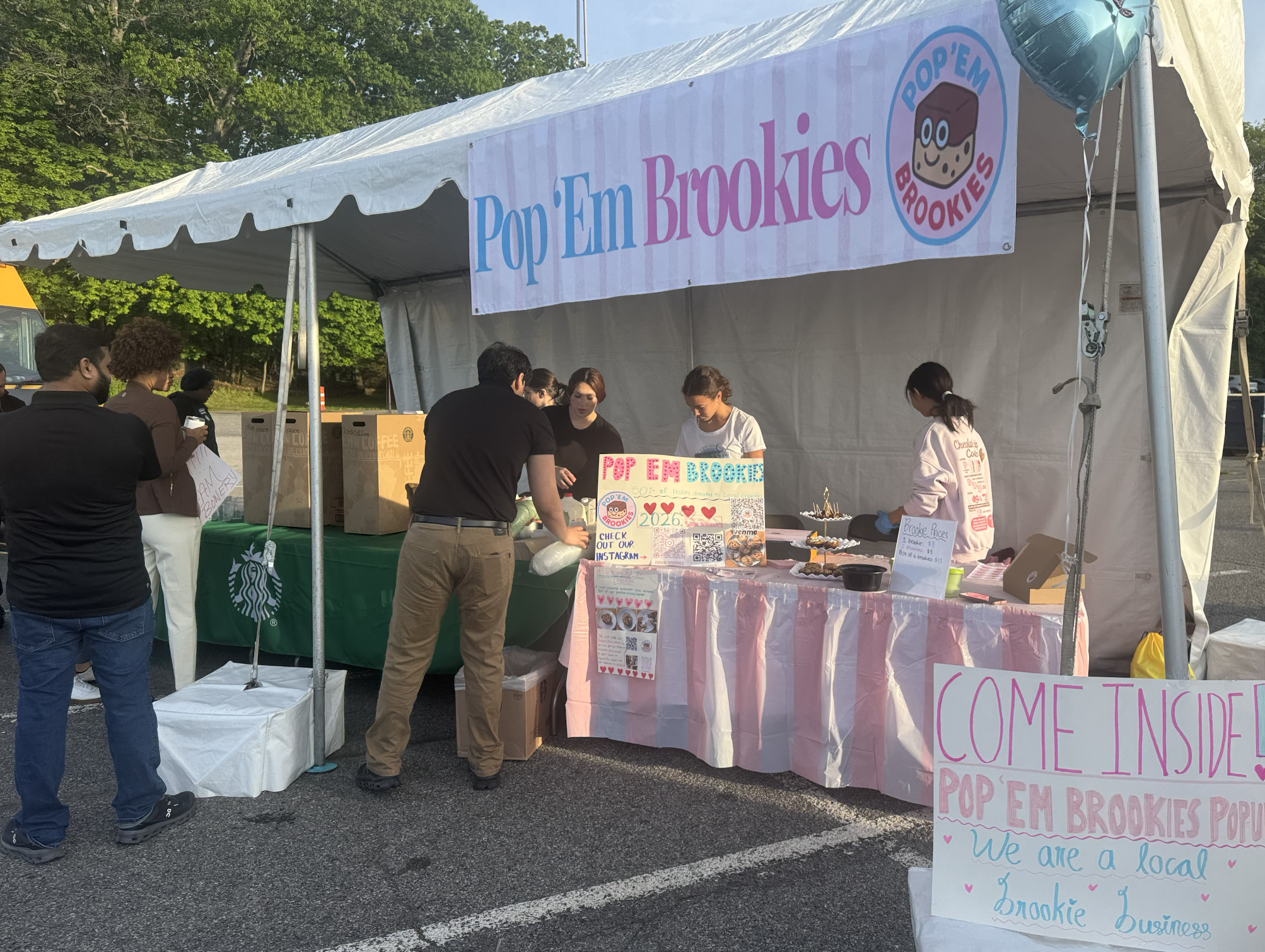 A food stand at an outdoor event with a large pink and blue sign that reads 'Pop 'Em Brookies' and a circular logo featuring a cookie dough brownie with eyes and a smile. Several people are gathered around the stand, ordering and selling baked goods. The stand has a pink tablecloth, and various signs with handwritten messages and prices are displayed.