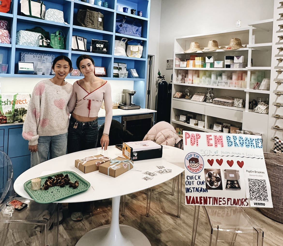 Two women standing behind a table with boxes of baked goods at a Valentine-themed bake sale, with a colorful sign promoting Pop 'Em Brookies and local fundraising.