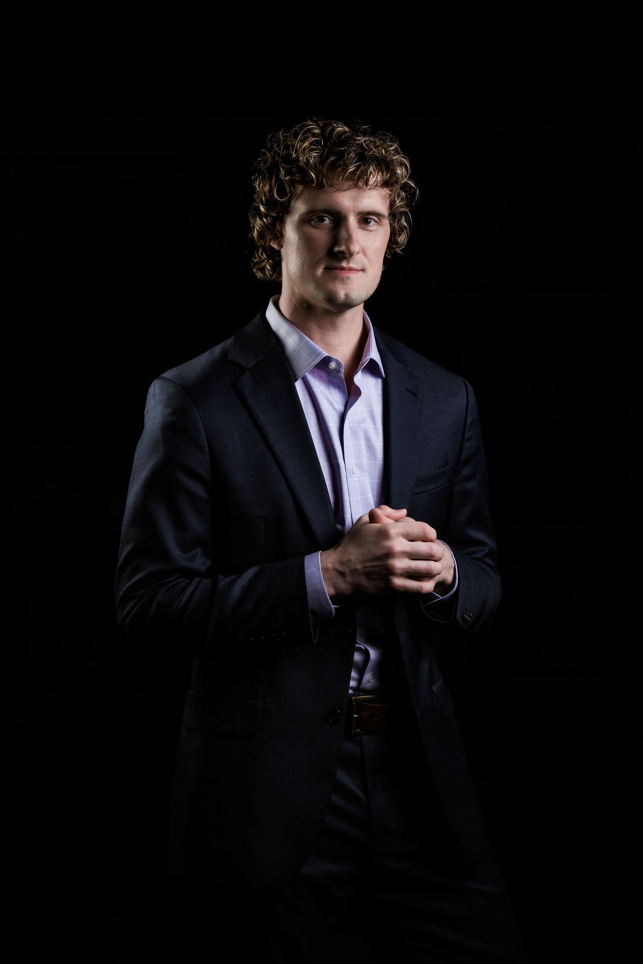 A man, Justin T. Weller (Justin Weller), with curly hair wearing a dark suit jacket and a light purple dress shirt, standing against a black background, looking confidently at the camera with hands clasped.
