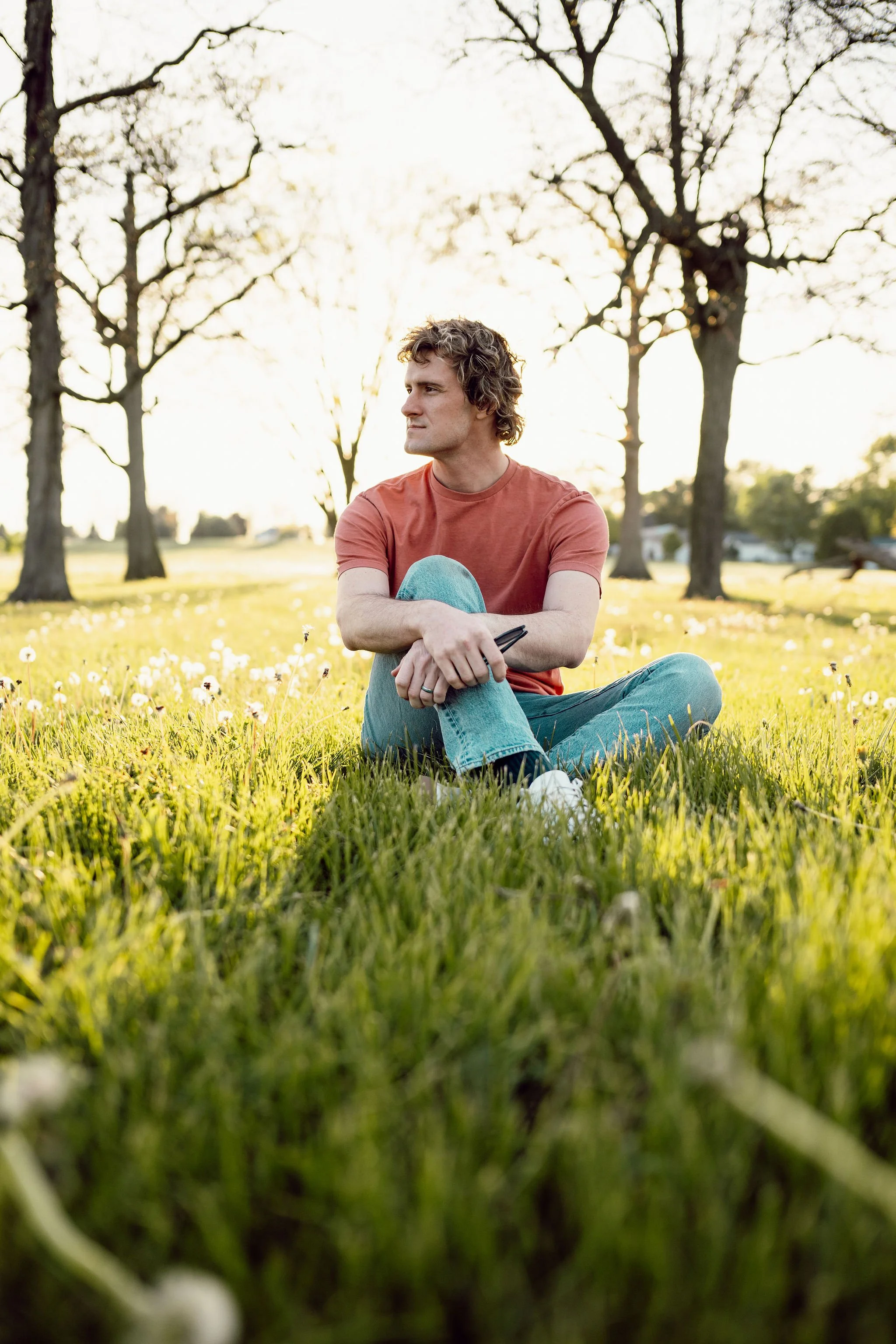 Justin T. Weller (Justin Weller) sits a grass field with trees and the sunset at his back while thoughtfully looking on.
