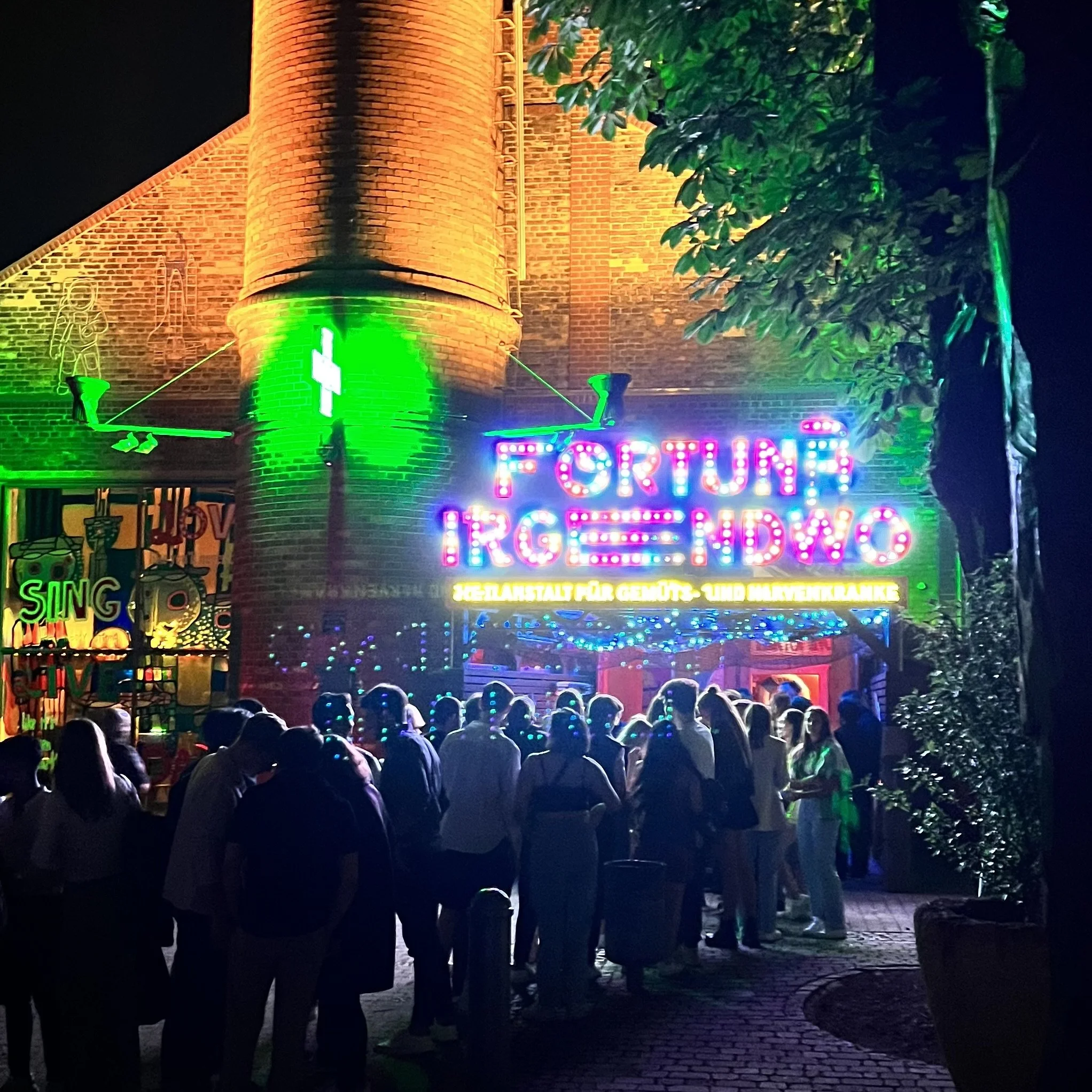 Besucher bei der Veranstaltung „Friends House“ im Fortuna Irgendwo in Frankfurt am Main, vor dem beleuchteten Turm und bunten Neonlichtern bei Nacht.