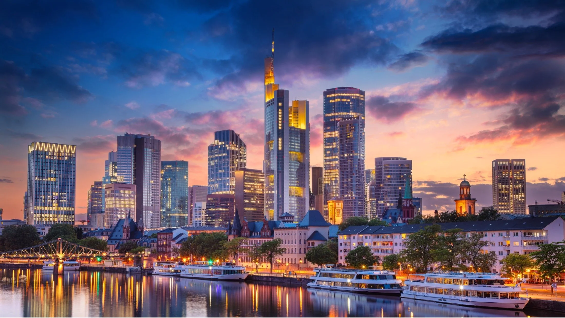 Stadtbild mit modernen Wolkenkratzern bei Sonnenuntergang, reflektiert im Fluss, mit Booten im Vordergrund.