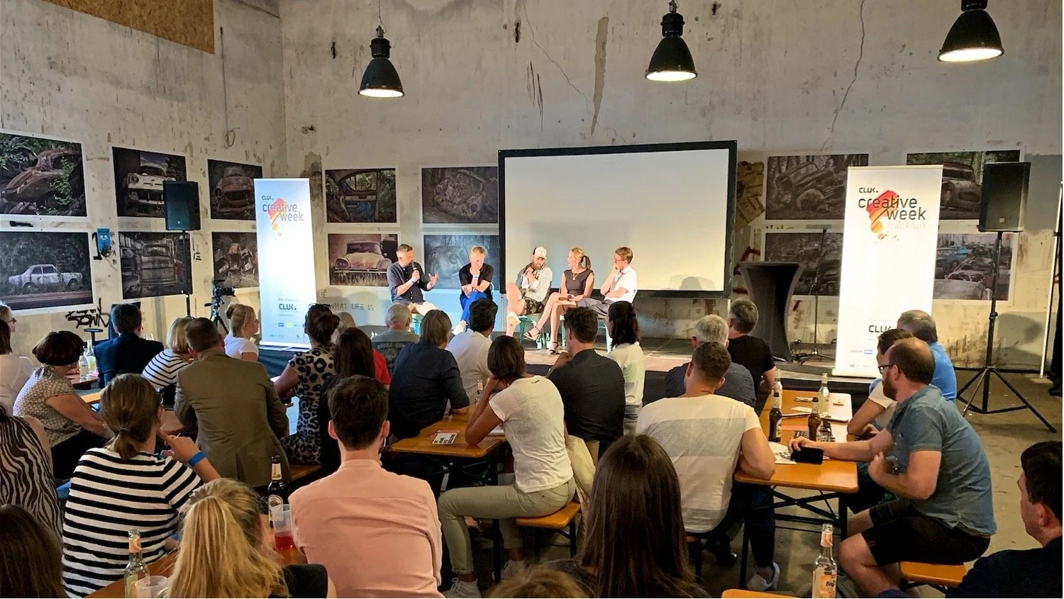 Eine Gruppe von Menschen sitzt an Tischen in einem Raum, der eine Podiumsdiskussion mit fünf Personen auf der Bühne zeigt. Die Bühne hat einen großen Bildschirm im Hintergrund und zwei gestreifte Banner mit dem Logo "creative week".