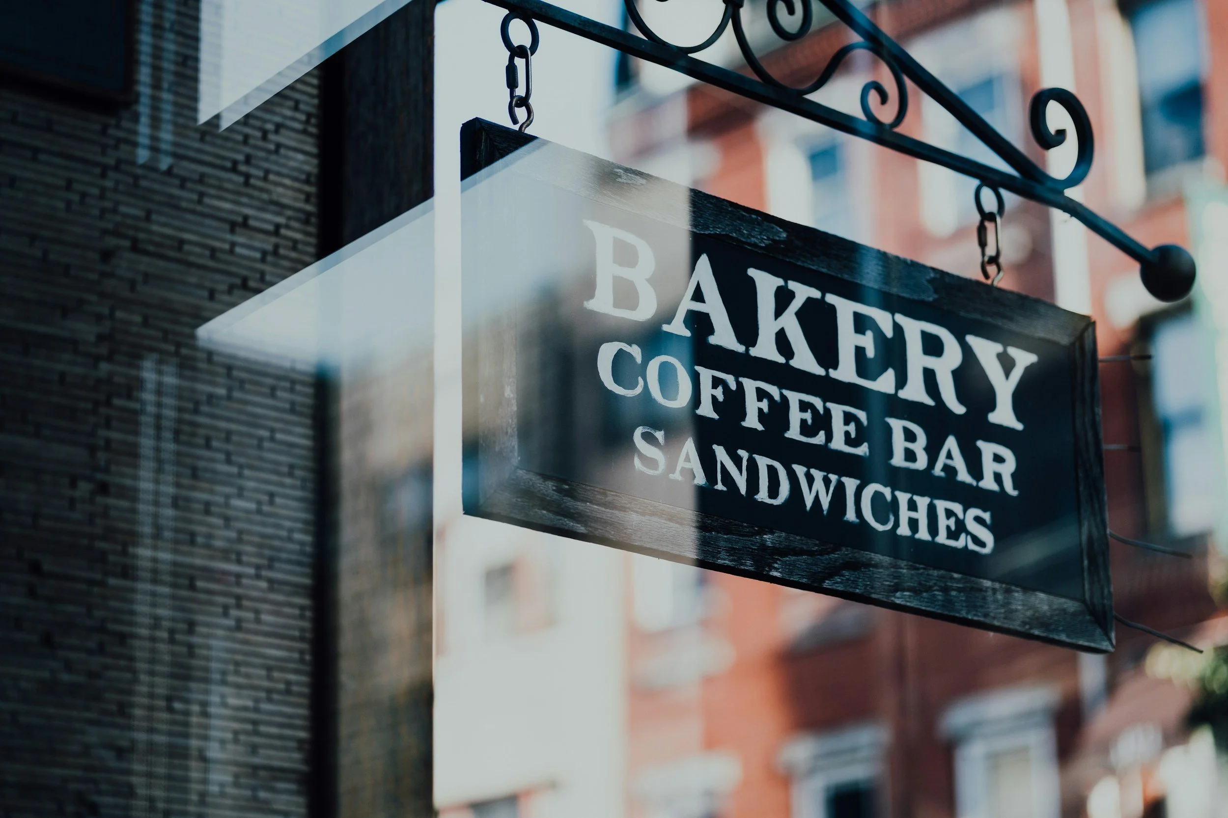 A blackboard sign hanging outside a bakery, coffee bar, and sandwich shop with the words 'Bakery,' 'Coffee Bar,' and 'Sandwiches' written on it.