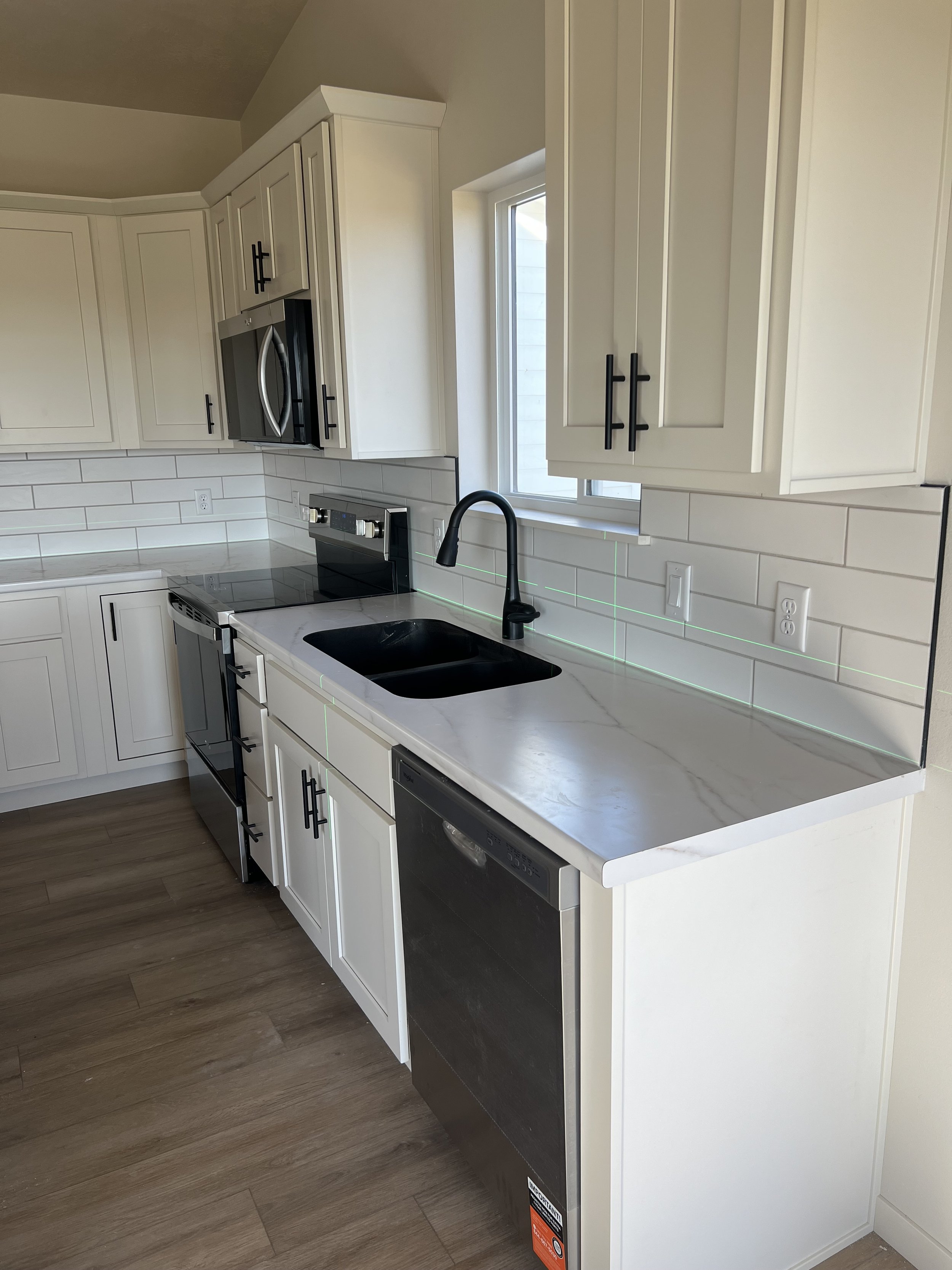 White kitchen cabinets, black hardware, black microwave oven, electric stove, white countertop, black sink, black faucet, window, white subway tile backsplash, electrical outlets, dishwasher, and wood flooring.