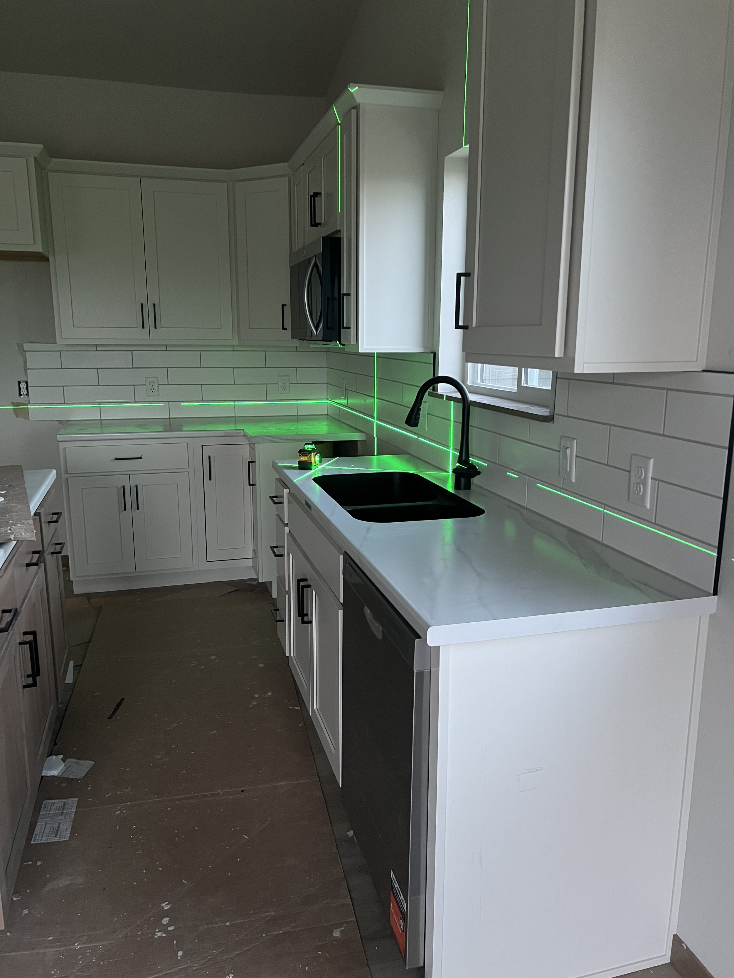 Modern kitchen with white cabinets, black handles, white tile backsplash, black sink faucet, and green laser lines outlining cabinet and wall edges, with a window on the right side.