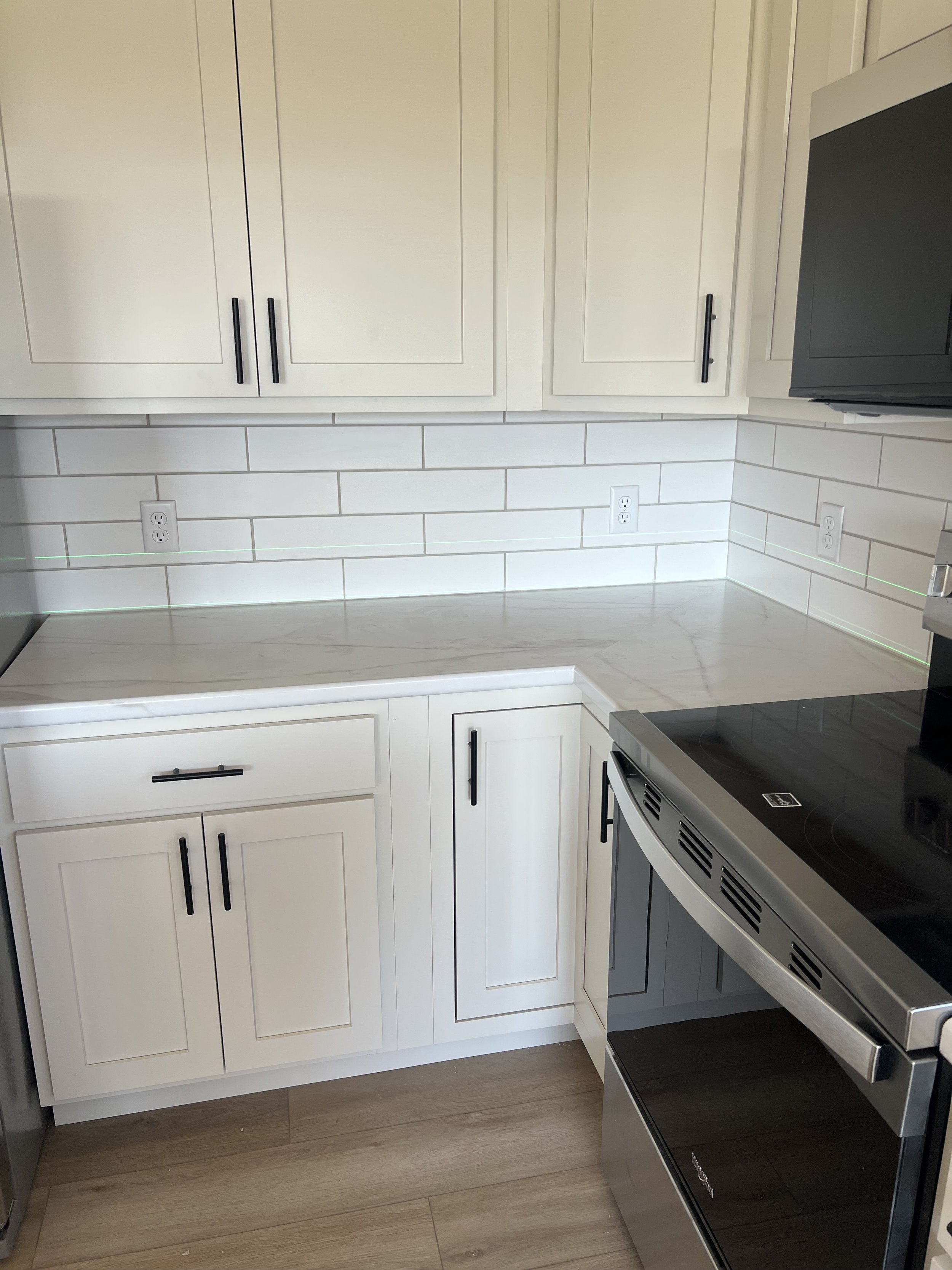 Modern kitchen with white cabinets, black handles, white subway tile backsplash, white countertop, stainless steel oven, and light wood flooring.