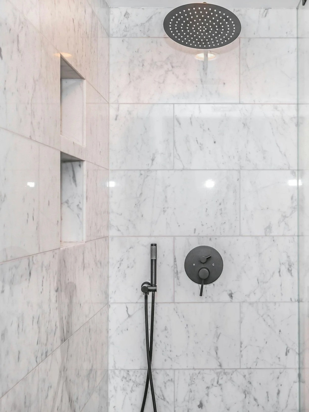 Bathroom shower with white marble tile walls, a round overhead shower head, a hand-held shower wand, and a temperature control knob.