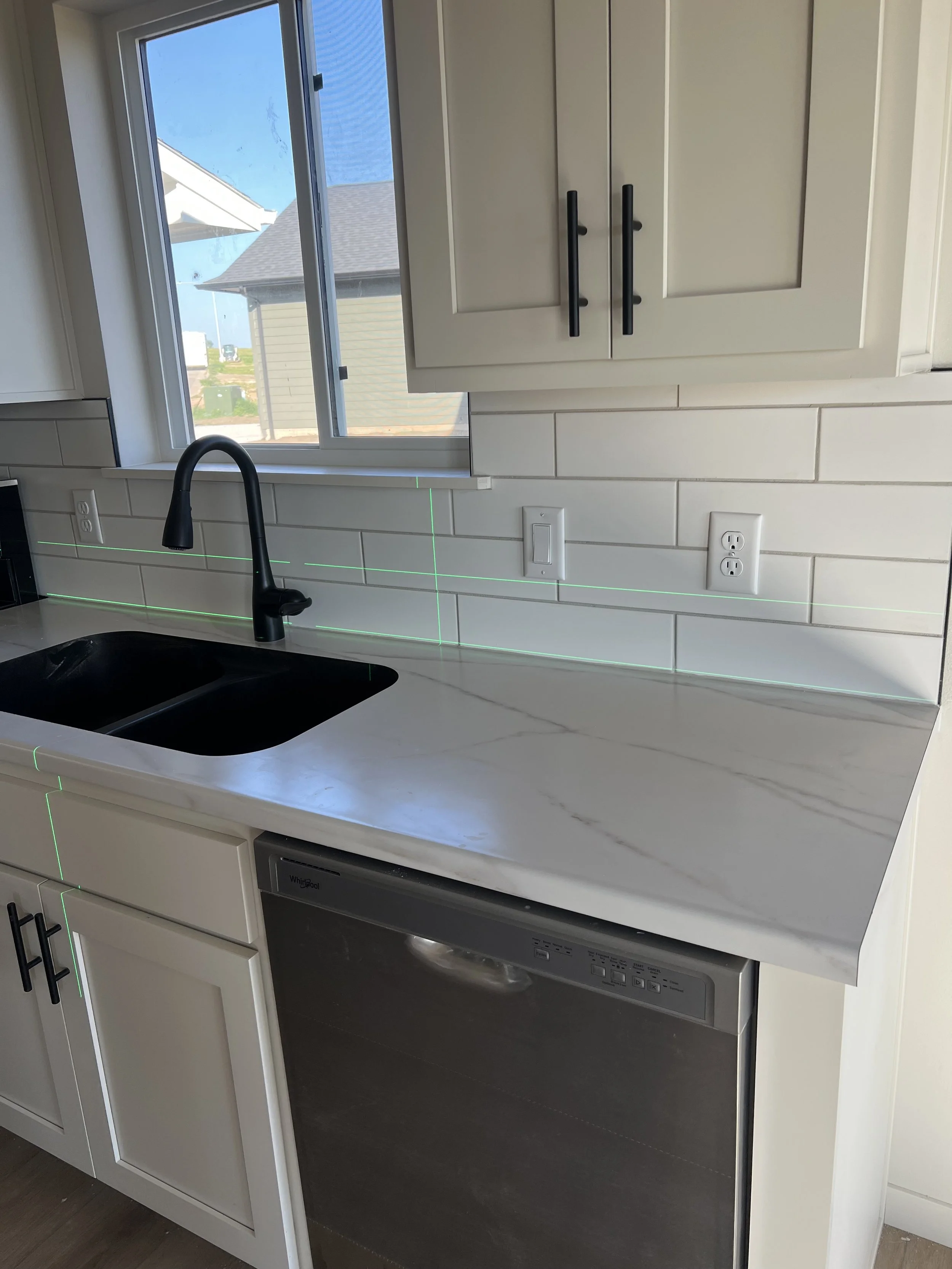 Modern kitchen with white cabinets, a black sink faucet, white marble countertop, tile backsplash, and a dishwasher. There is a window above the counter showing neighboring houses and a blue sky.