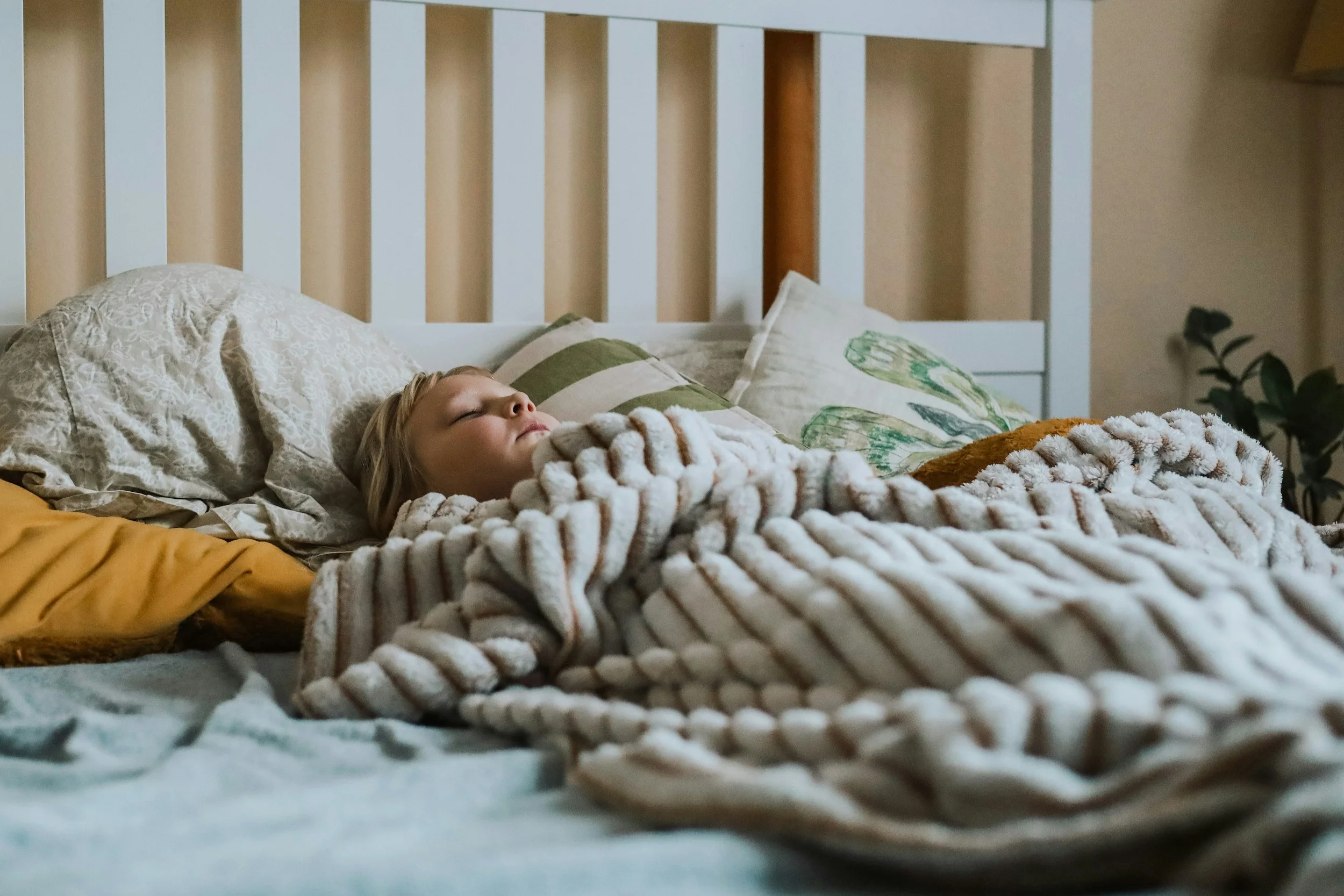 A young girl with blonde hair sleeping peacefully in bed, covered with a chunky knitted blanket, with pillows behind her, in a cozy bedroom setting.
