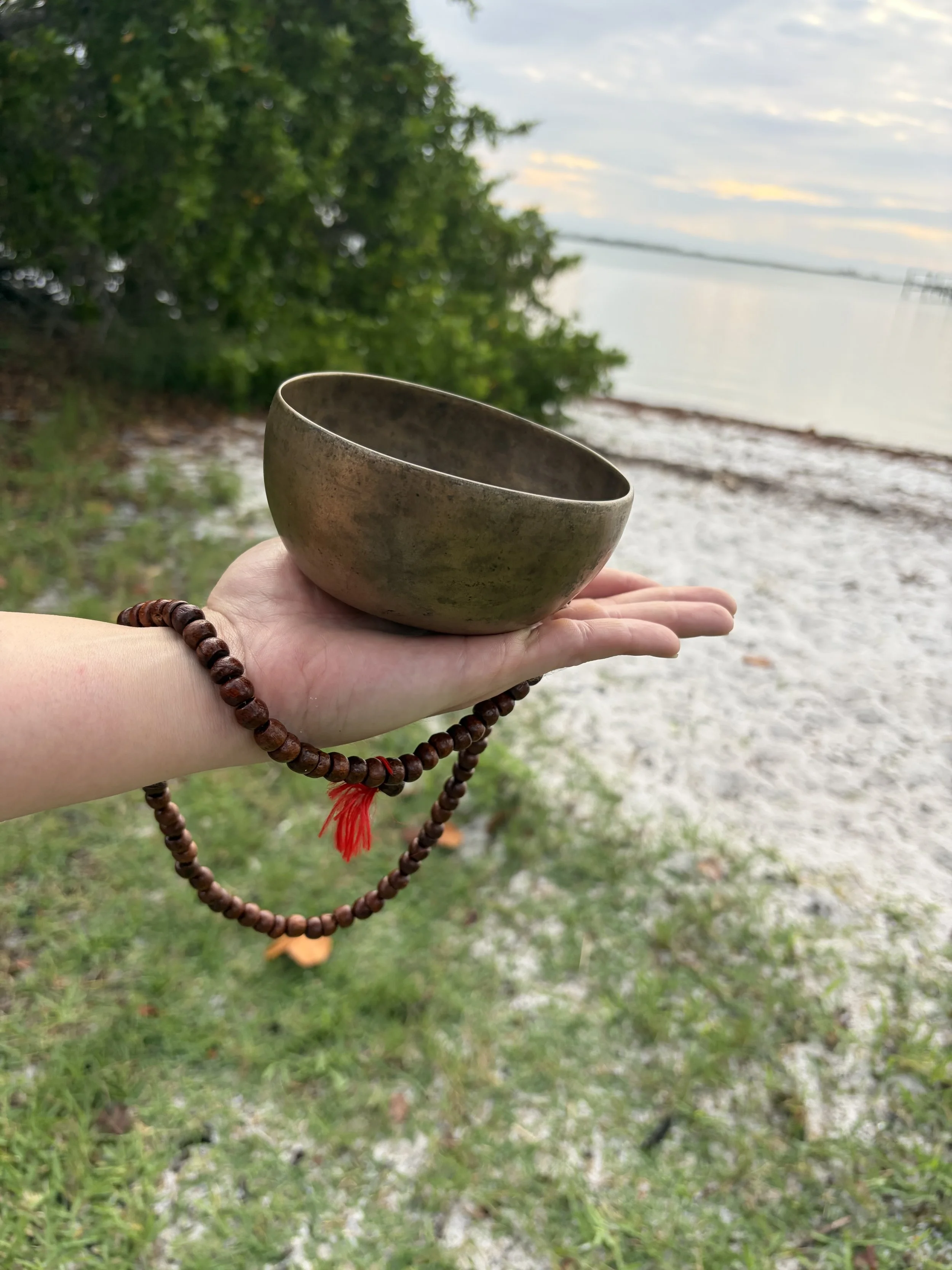 A person holding a small ceramic bowl with a red string of prayer beads around their wrist at a beachside location with trees, water, and a cloudy sky in the background.