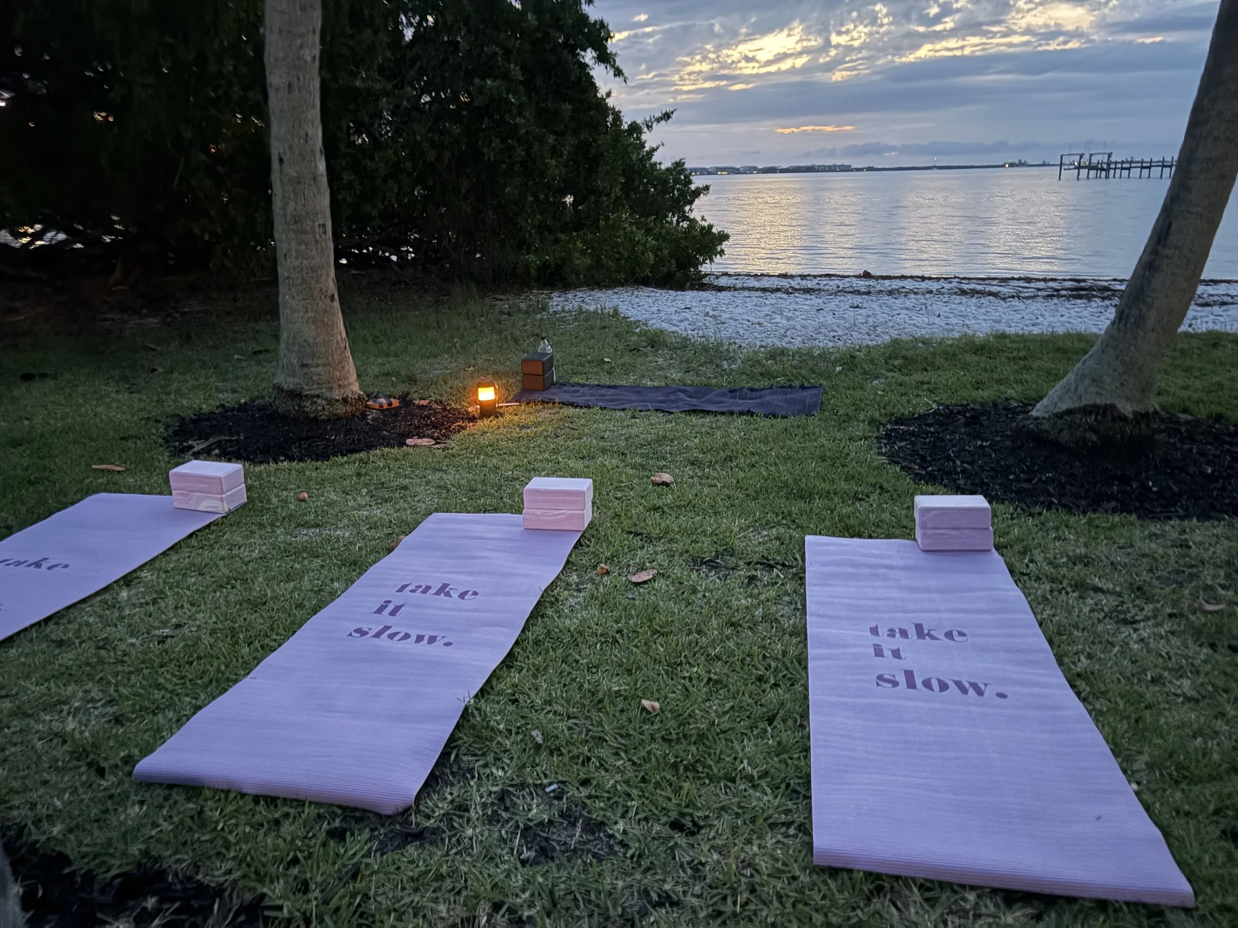 An outdoor setting by a lake at dusk with yoga mats on the grass, each labeled with the words 'lake it slow,' and small lanterns providing light. In the background, the lake and sky are visible with some clouds and distant structures.