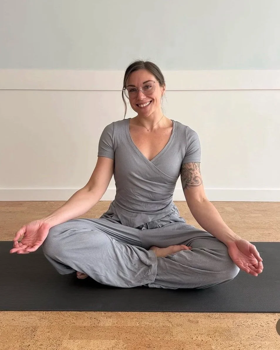 Woman sitting cross-legged on yoga mat, smiling, in a light-colored room, practicing mindfulness or yoga.