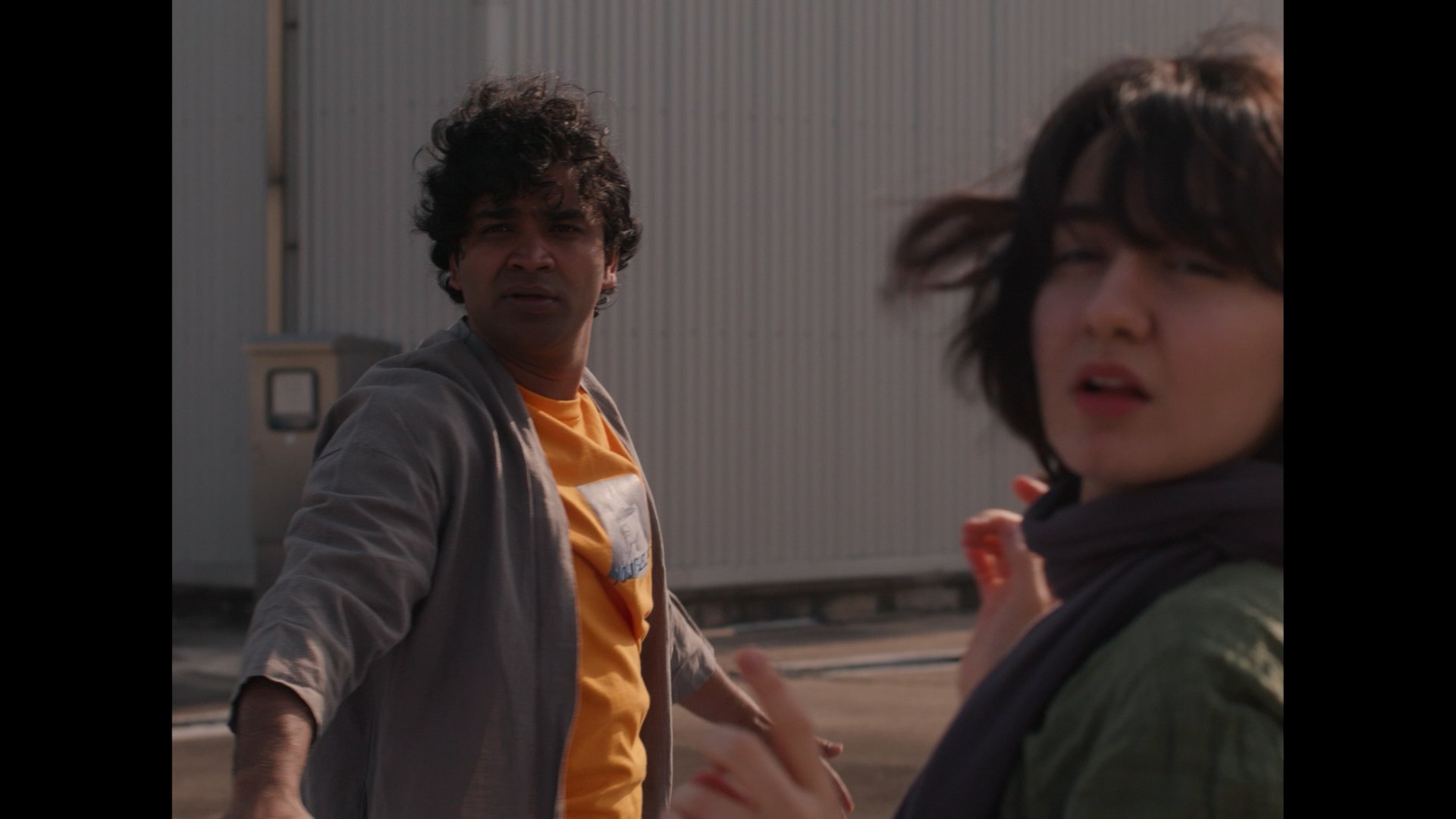 Two people standing outdoors near a corrugated metal building, a man with curly hair wearing a yellow shirt and a woman with short hair wearing a jacket engage in conversation.