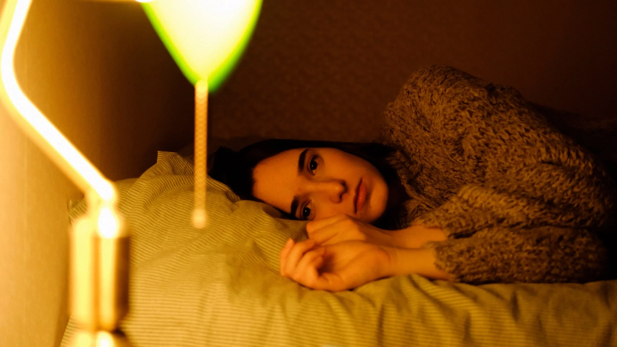 A woman lying on a bed, resting her head on a striped pillow, looking at a candle with a focused expression, wearing a knitted sweater in a dimly lit room.