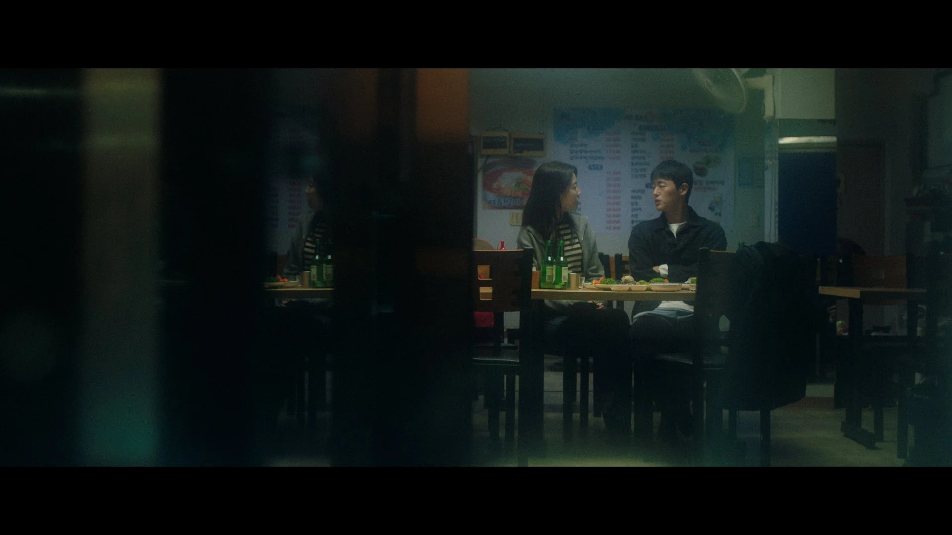 A scene inside a dimly lit restaurant with two people sitting at a table and talking, seen through a partially obscured view.