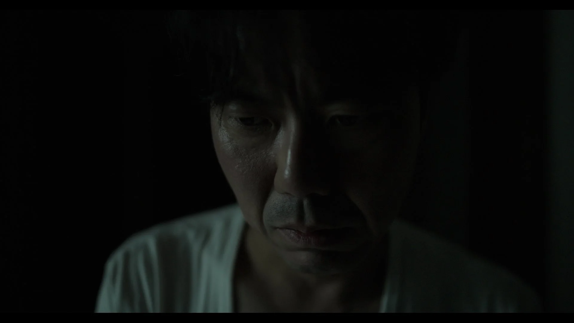 Close-up of a man's face in darkness, with only part of his face illuminated, showing a serious expression.