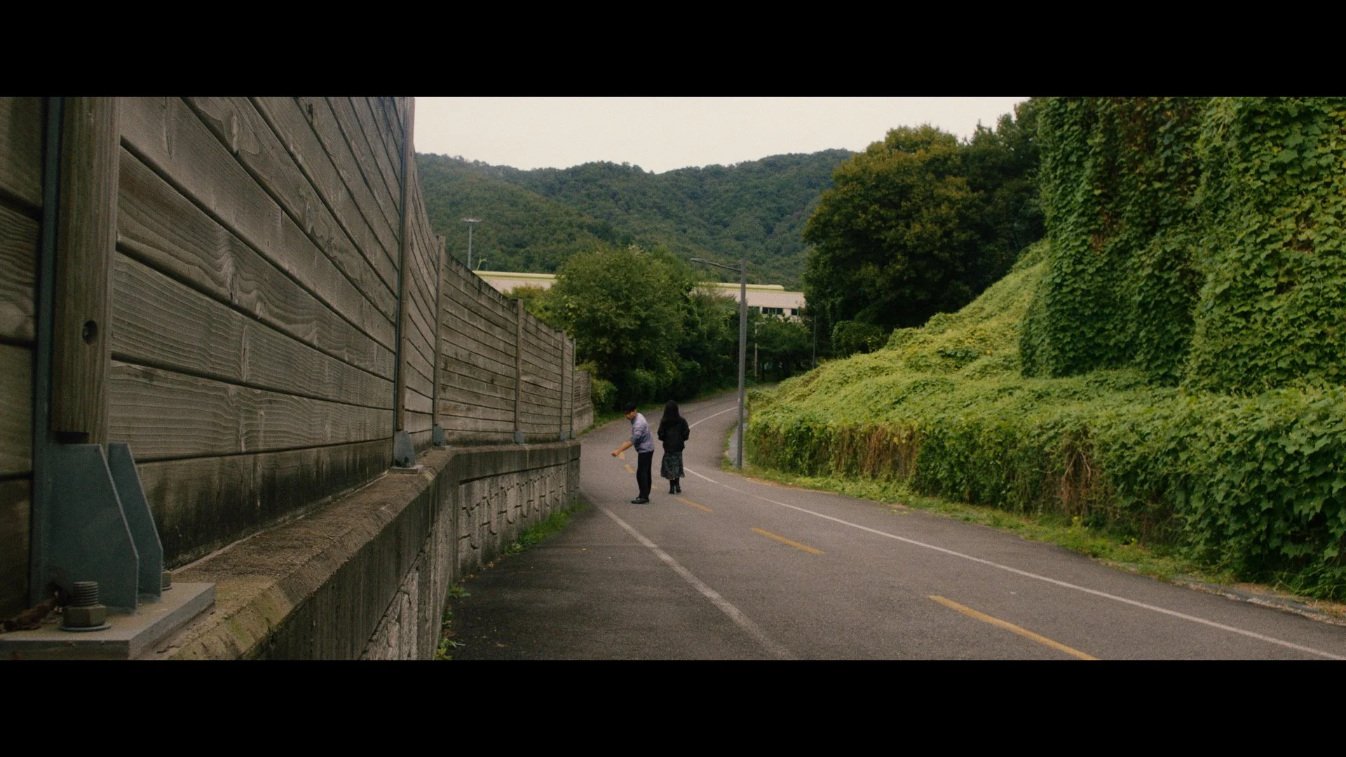 Two people walking along a winding mountain road with a wooden fence on the left and greenery on the right.