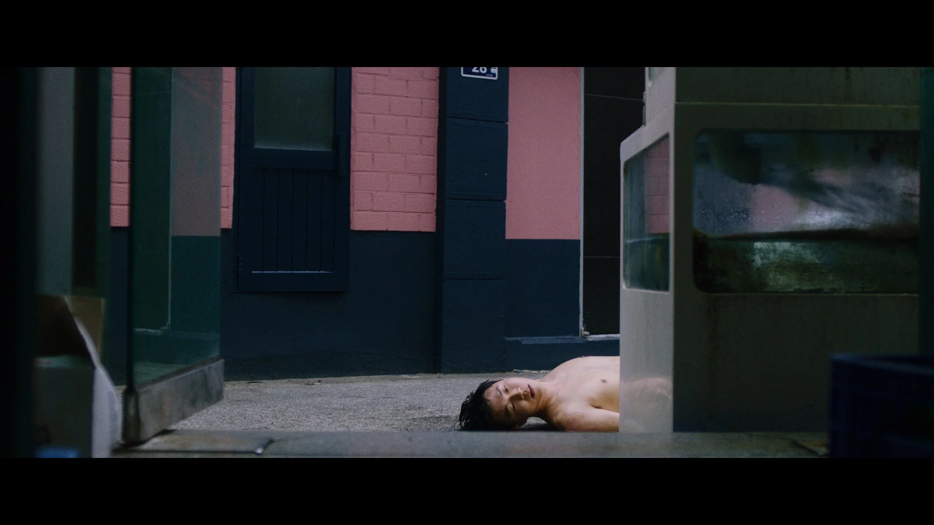 A woman lying on the ground topless outside a building with pink and dark blue walls, seen through a gap between a glass display case and another object.