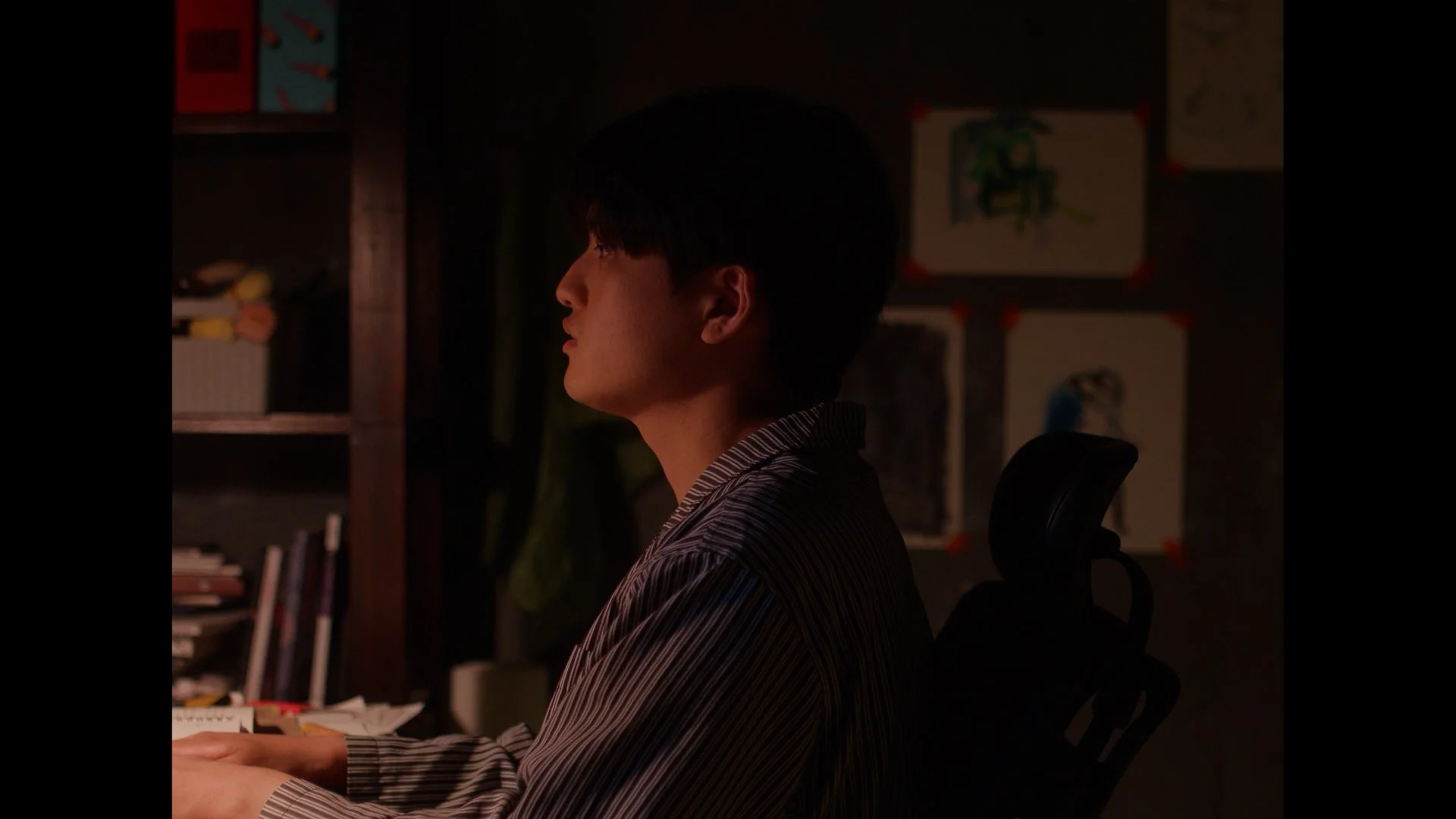 Side profile of a young man with dark hair sitting at a desk in a dimly lit room, with bookshelves and artwork in the background.