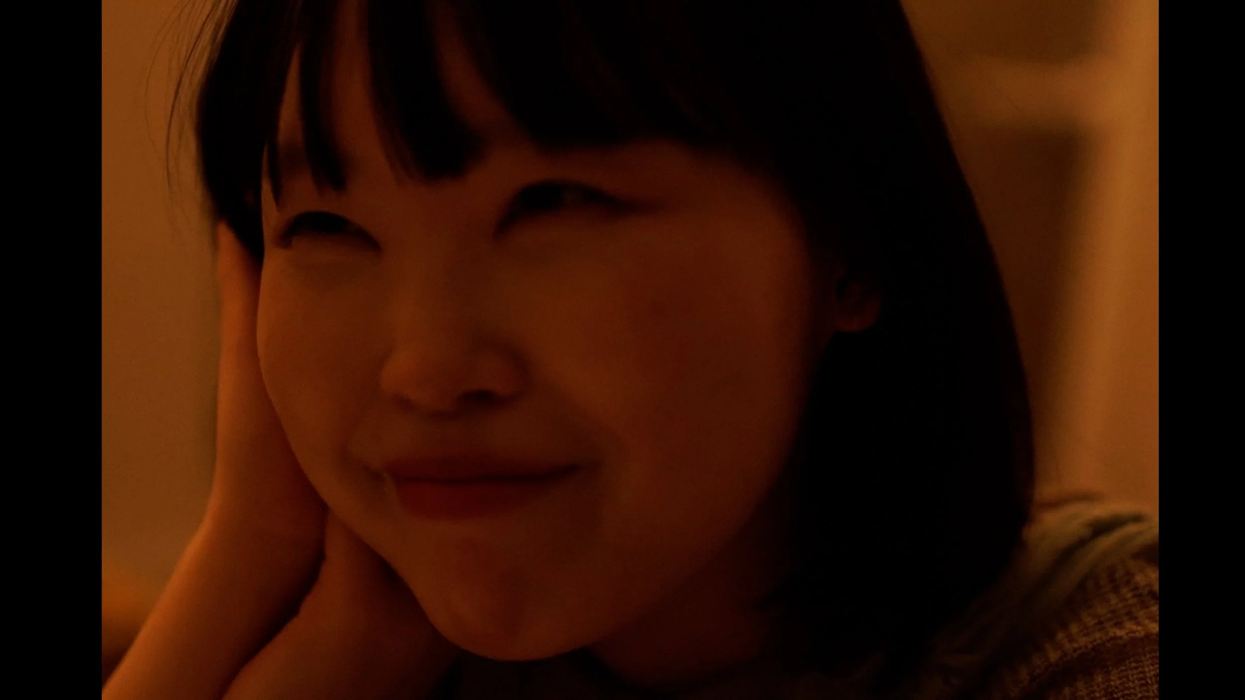 Close-up of a young woman smiling softly, resting her head on her hand with dark hair and bangs.