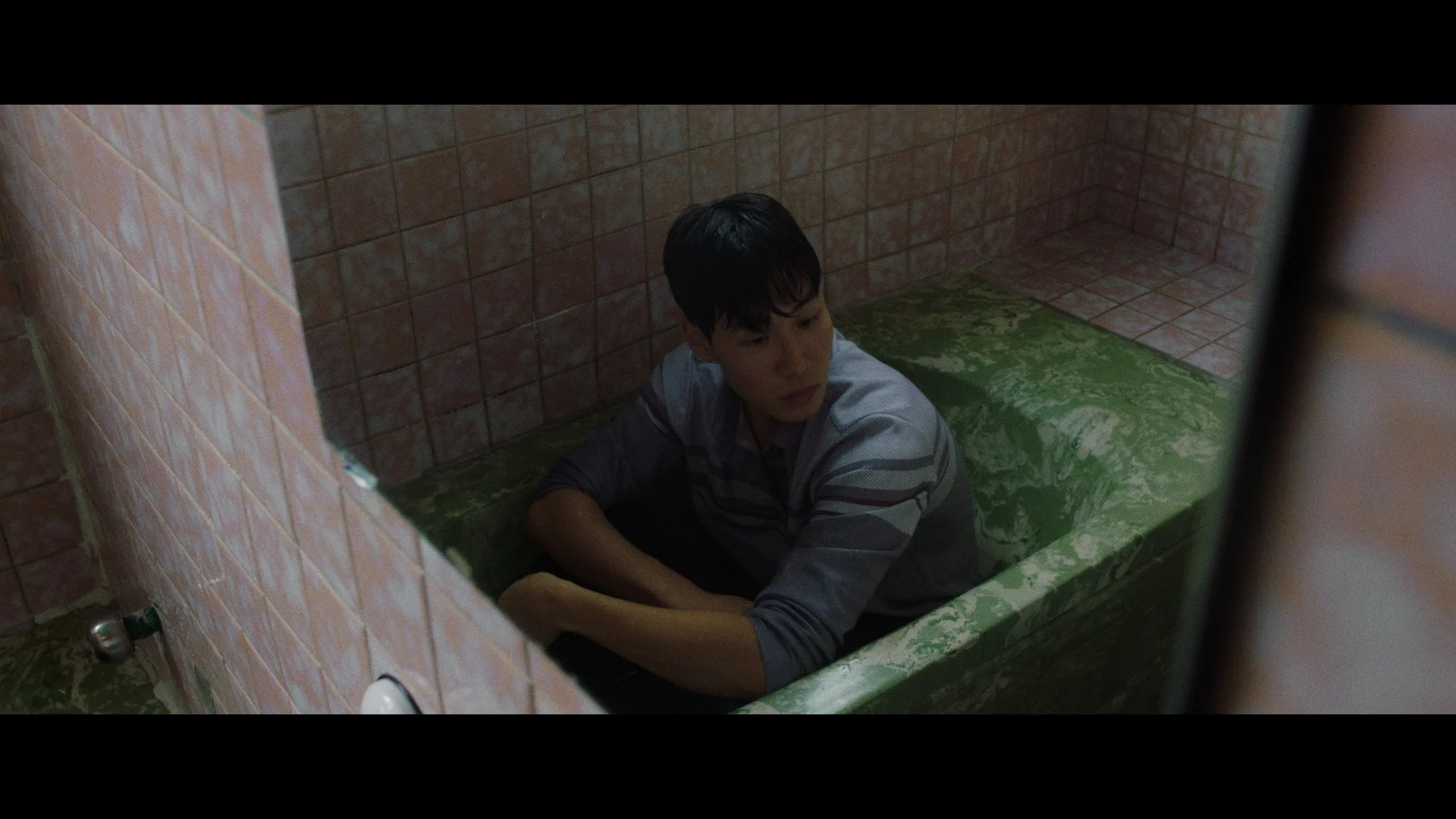 A young boy sitting in a green bathtub filled with water inside a tiled bathroom.